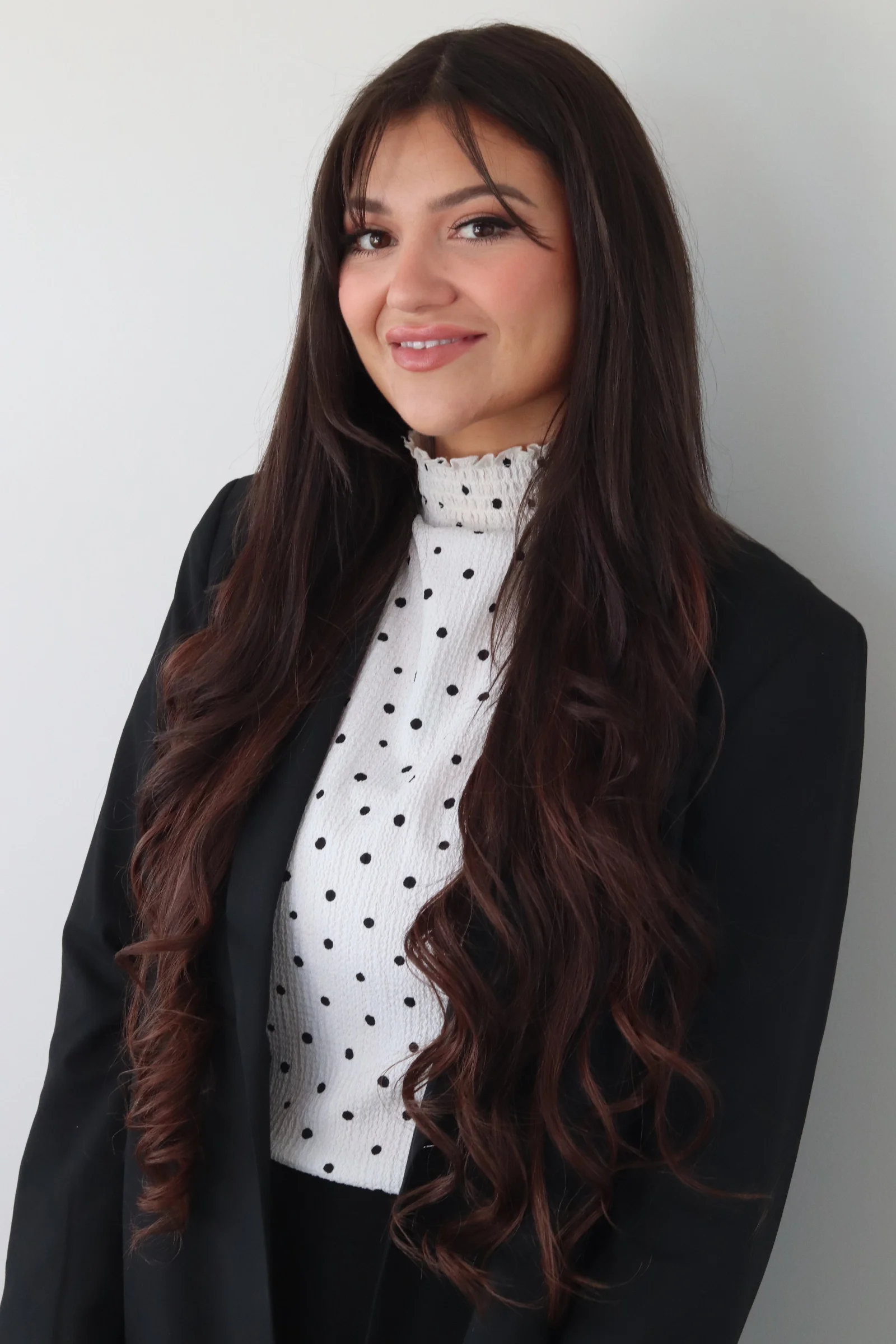 A woman with long, wavy brown hair and light skin, wearing a white turtleneck with black polka dots and a black blazer, smiling against a plain white background.