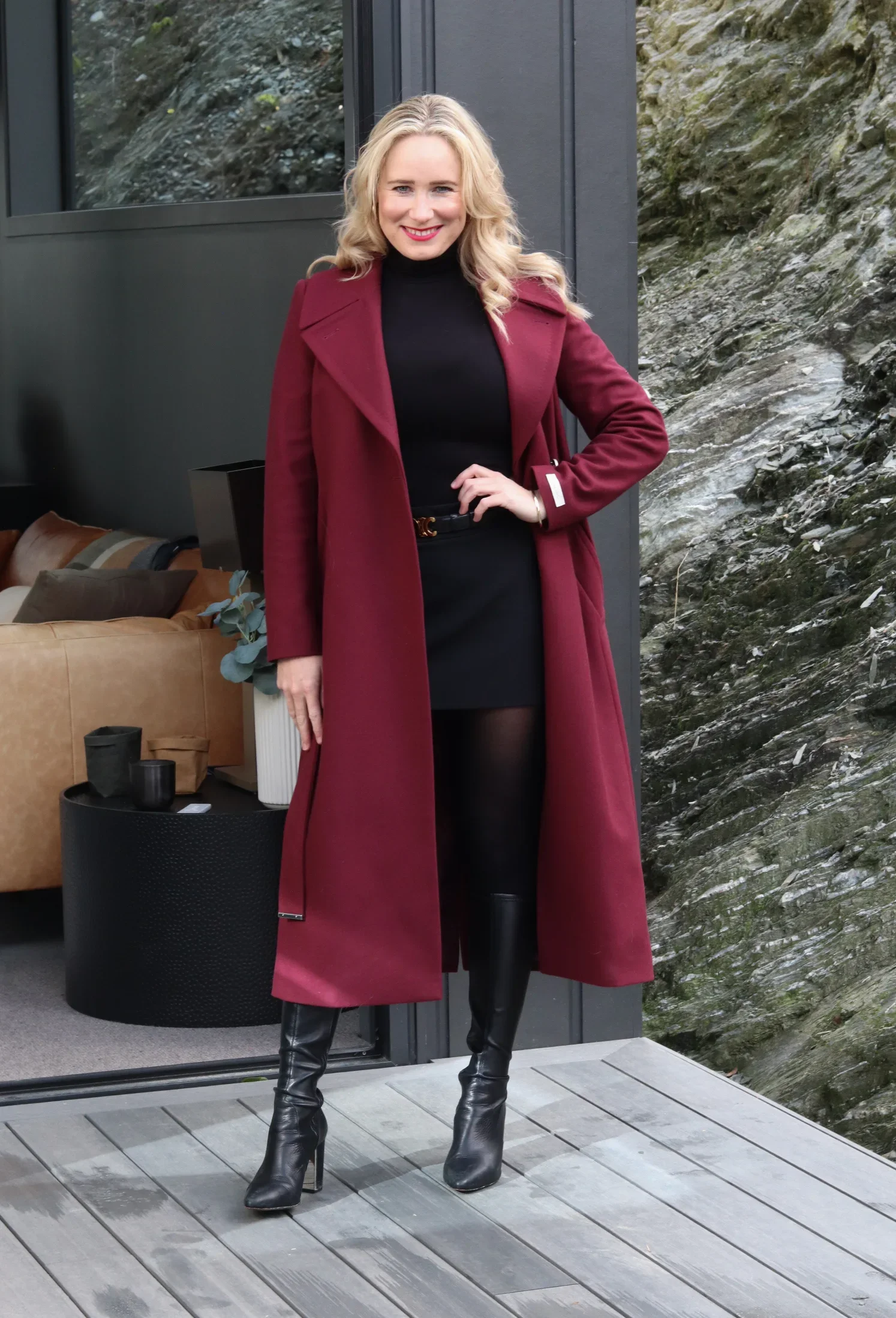 A woman with blonde curly hair smiling while standing on a porch, wearing a long burgundy coat, black turtleneck, black skirt, black tights, and black knee-high boots with heels.
