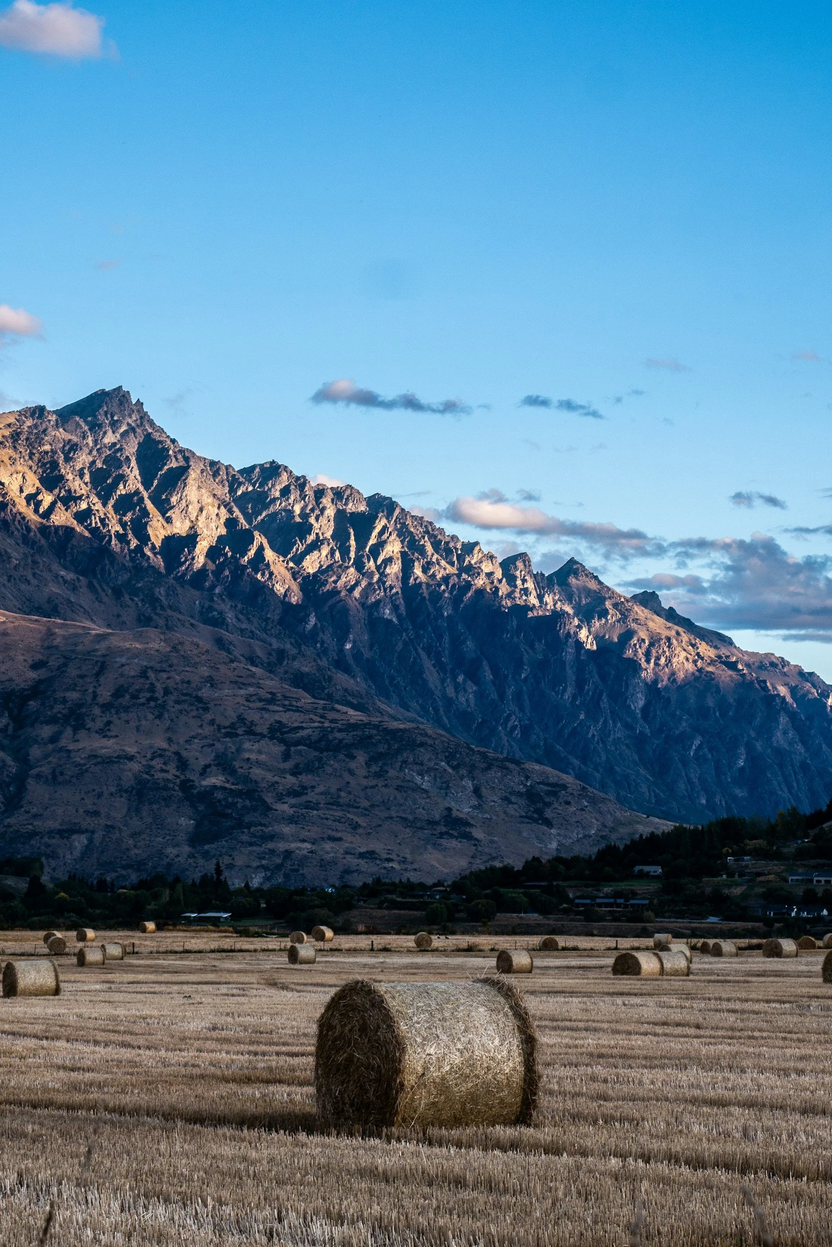 A rural field with hay bales, with mountain range and blue sky in the background under partly cloudy weather.