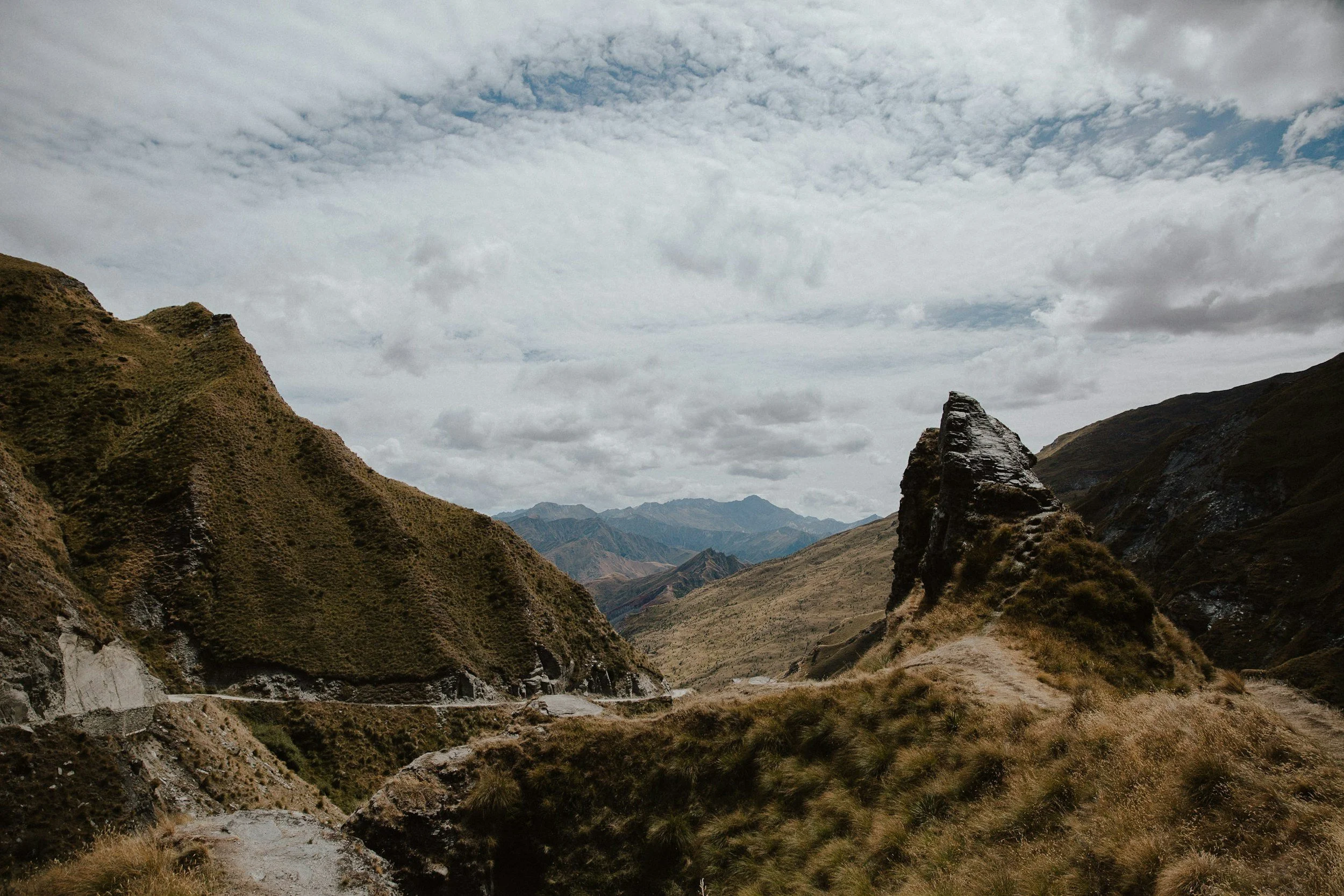 Mountain landscape with rugged hills, a dirt trail, and cloudy sky.
