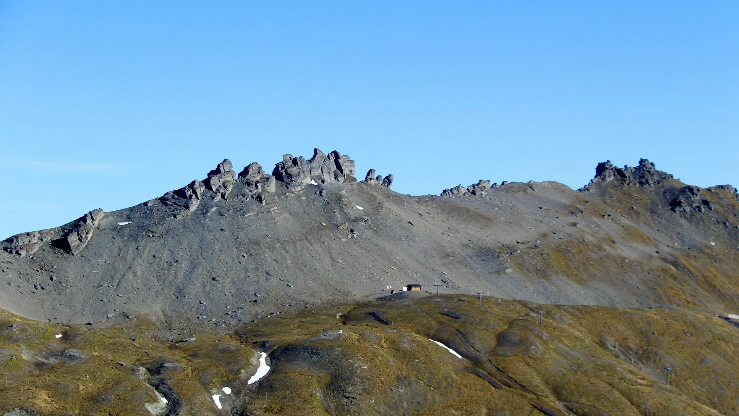 A rugged mountain landscape with steep rocky slopes and patches of snow, under a clear blue sky.