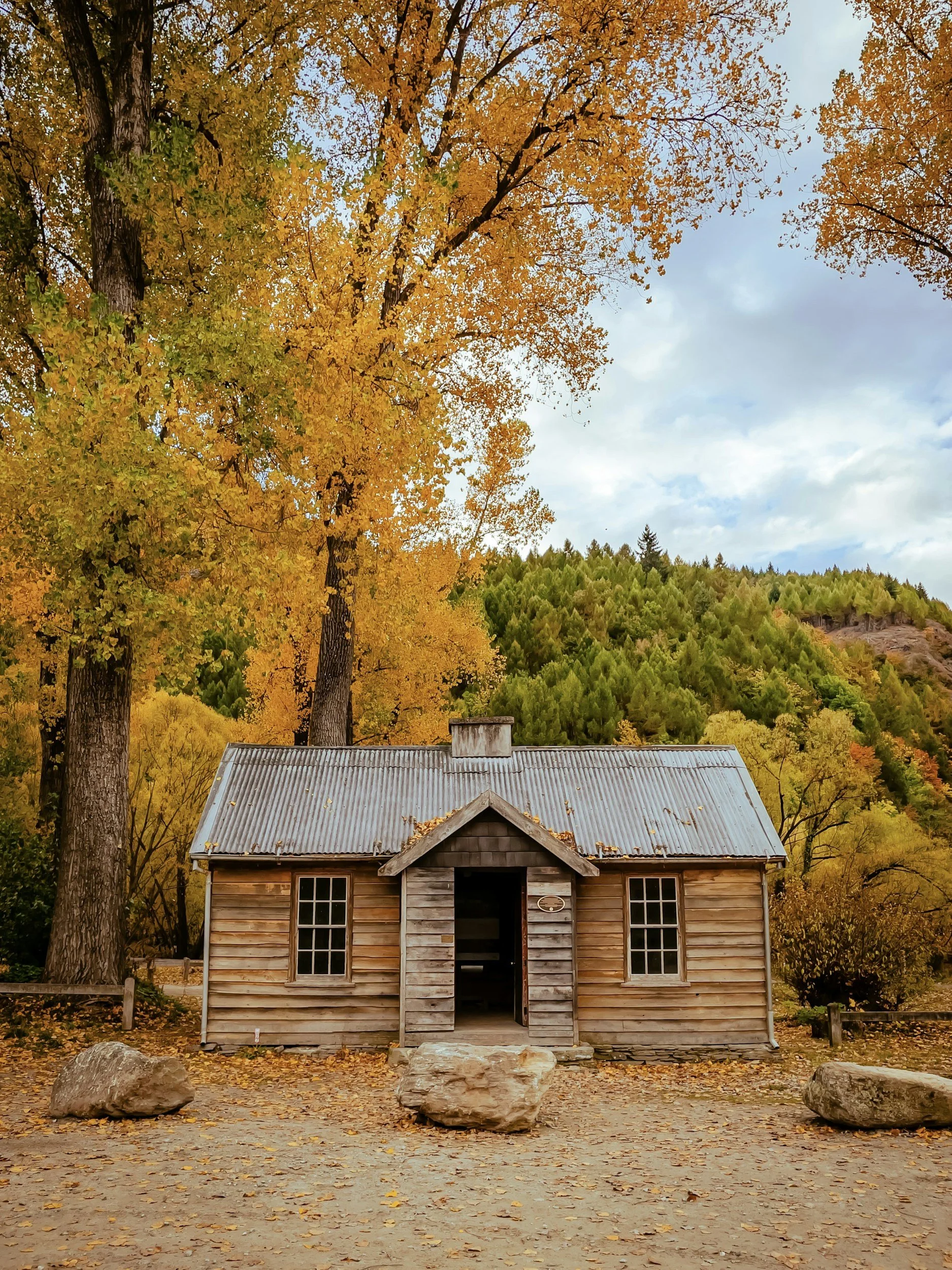 A small rustic wooden cabin with a metal roof, surrounded by fall trees with yellow and orange leaves, rocks, and fallen leaves on the ground.