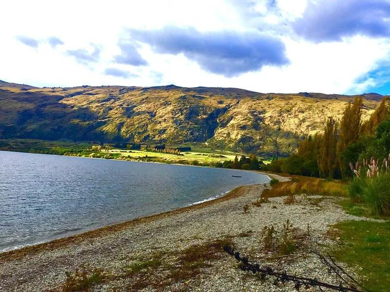 A lakeside shoreline with a mountain backdrop under a partly cloudy sky.
