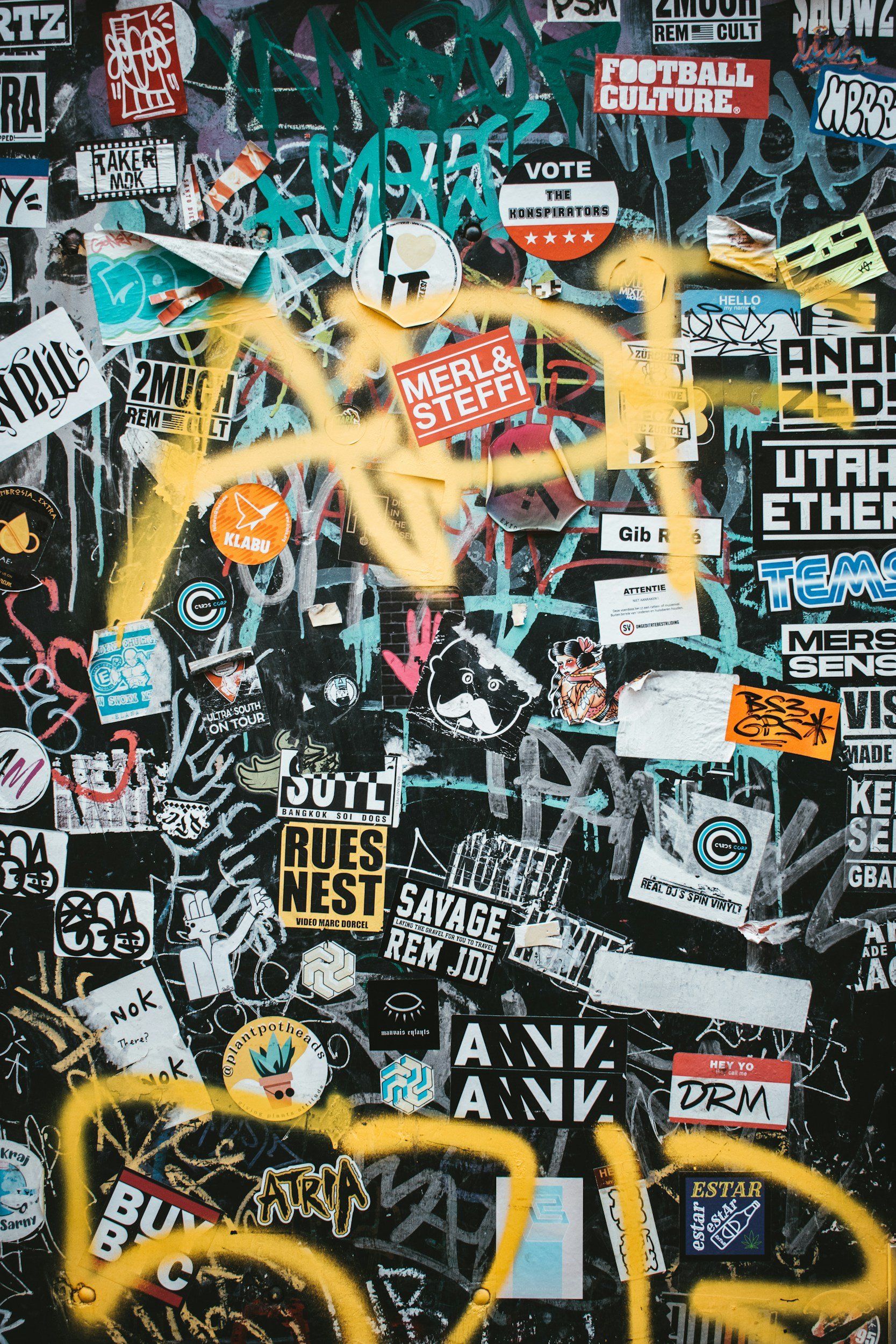 A wall covered with various graffiti stickers, posters, and street art, featuring different text, graphics, and tags with some colorful spray paint elements.