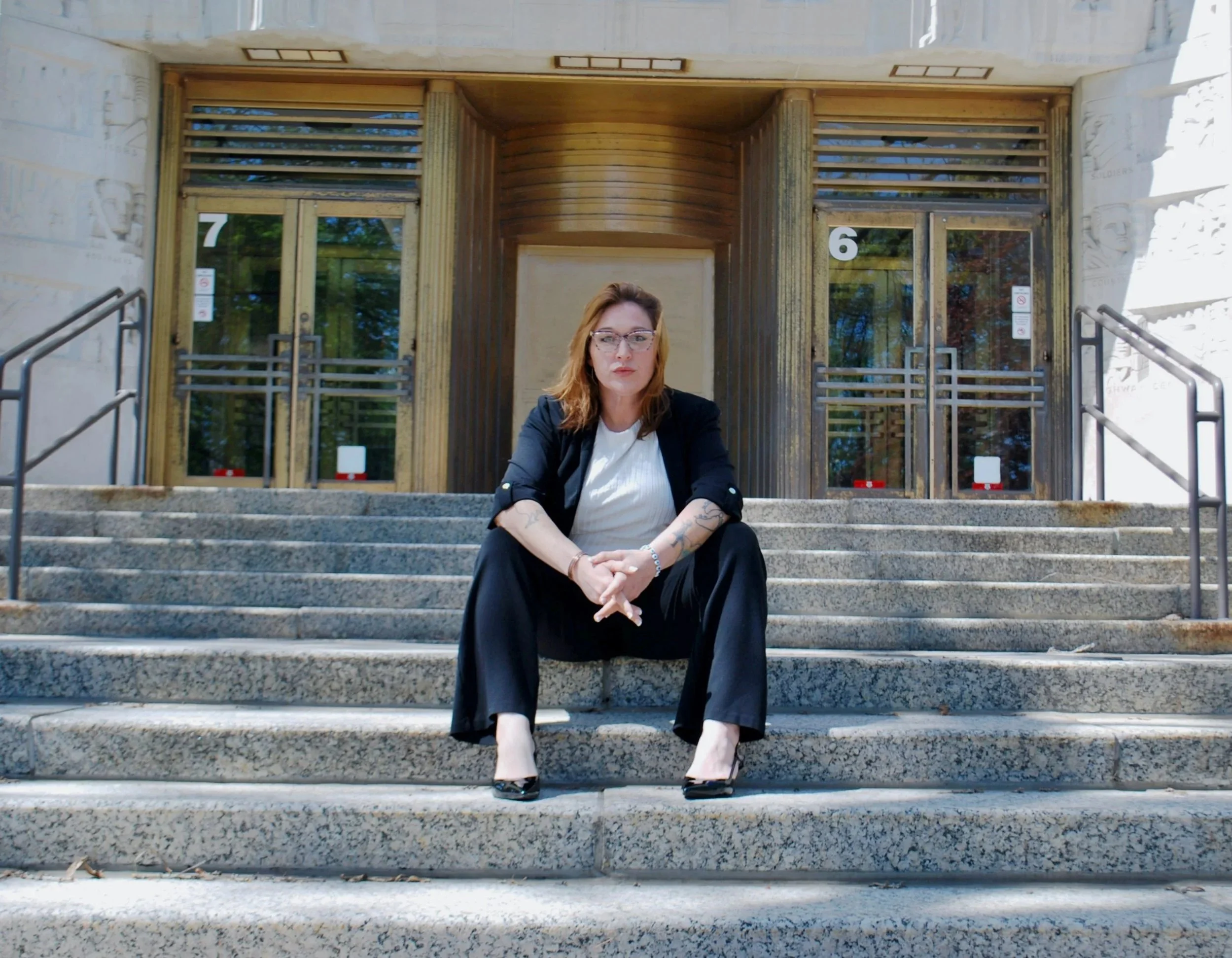 Woman with glasses sitting on stone steps in front of a building entrance with large glass doors, numbered 6 and 7, and architectural details on the sides.