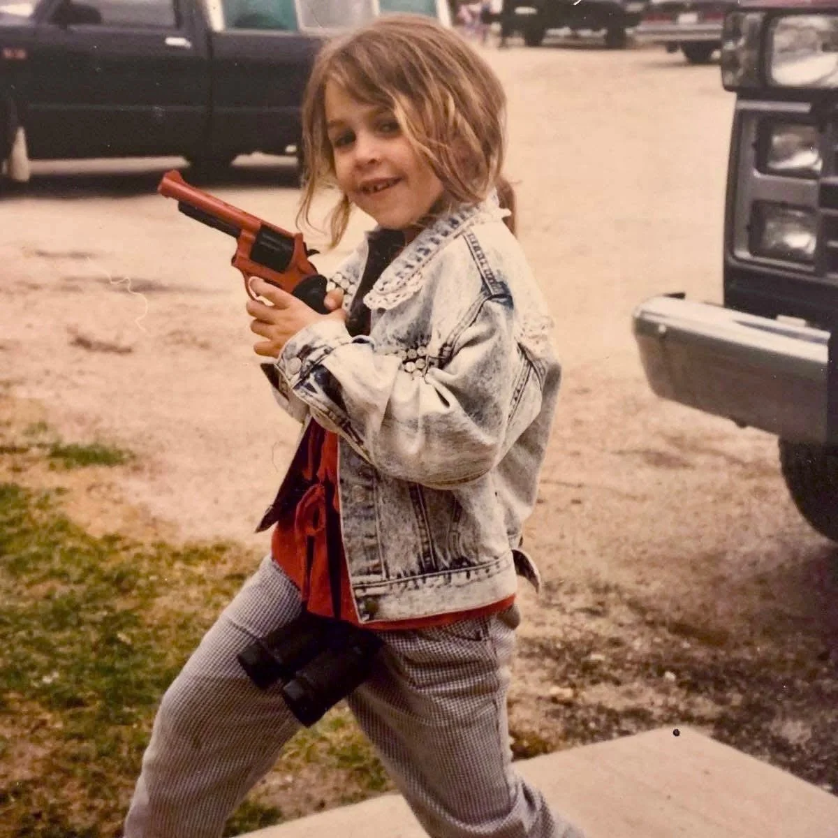 An image of Dani in 1989 with messy hair dressed in a denim jacket and checkered pants, holding a toy gun, standing outdoors near parked cars.