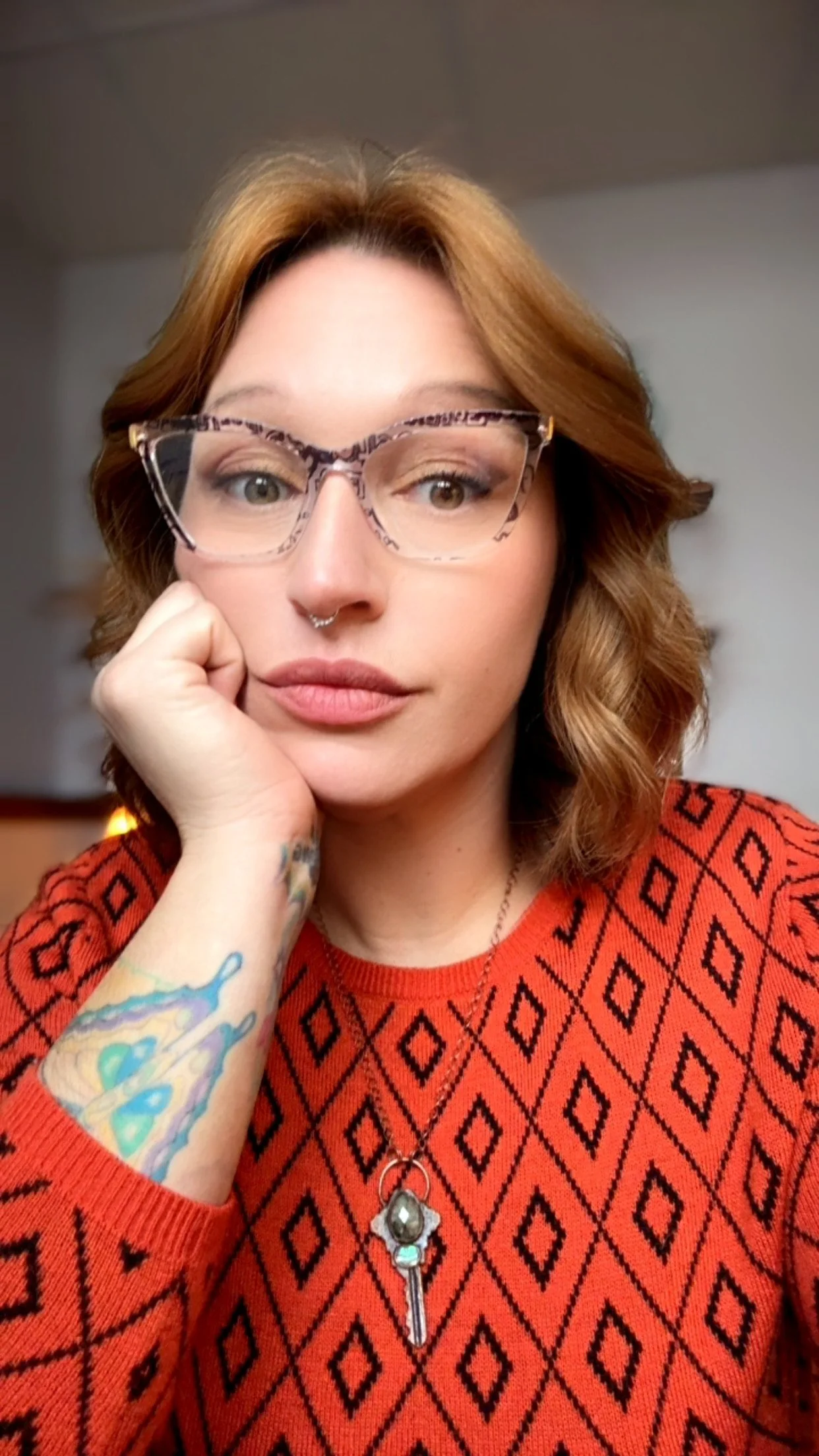 A woman with red hair, glasses, and a septum piercing wearing a red patterned sweater, resting her face on her hand, with tattoos on her arm, and a key-shaped necklace.