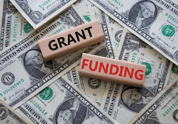 Two wooden blocks, one with the word 'GRANT' in black letters and the other with 'FUNDING' in red letters, placed on scattered U.S. dollar bills.