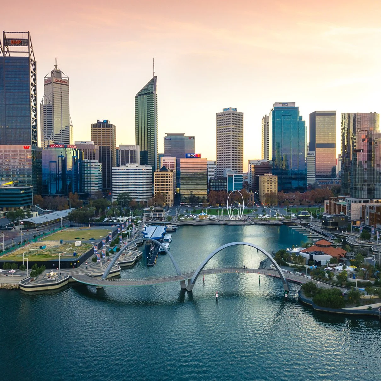 City skyline with tall modern skyscrapers, a waterfront park with bridges, sculptures, and recreational areas during sunset.