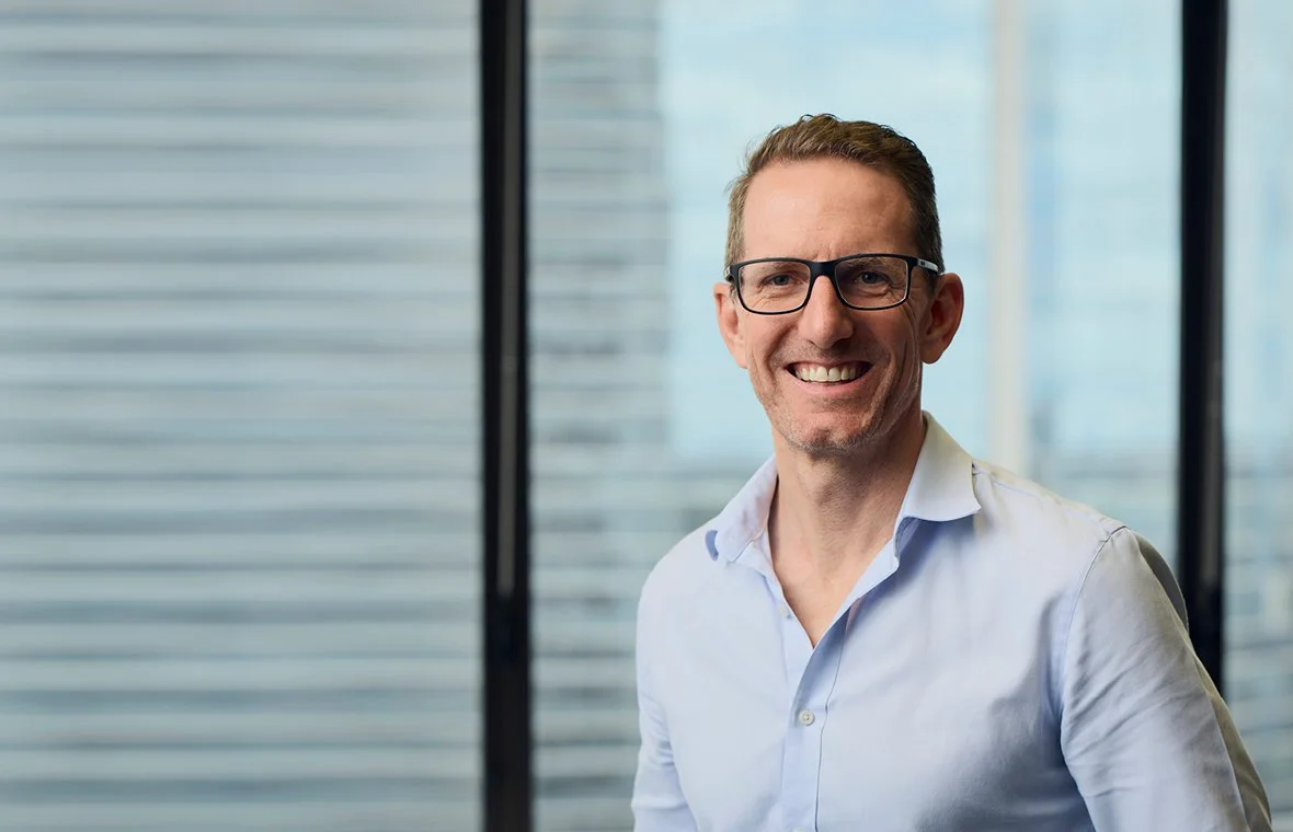 Smiling man with glasses in professional attire standing in front of office windows.