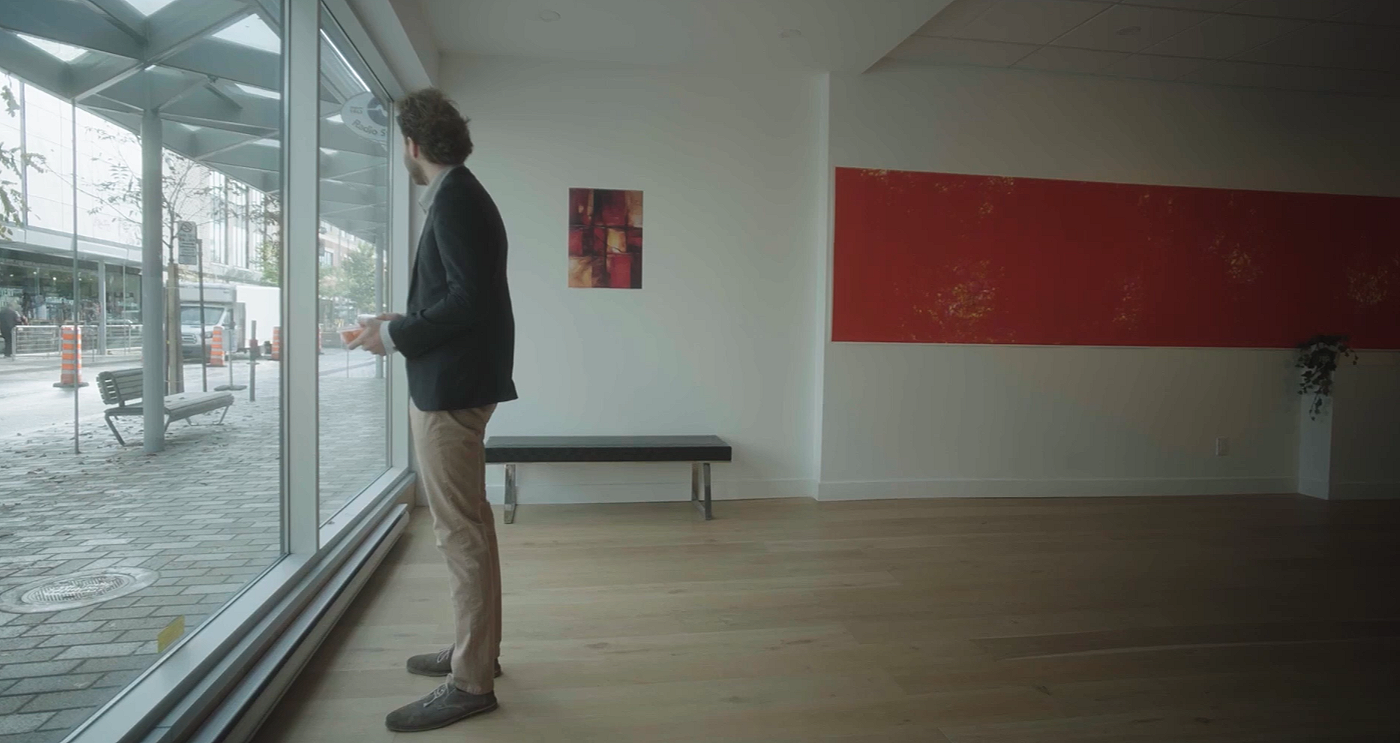 Un homme debout dans une salle presque vide regarde par une grande vitre vers la rue.