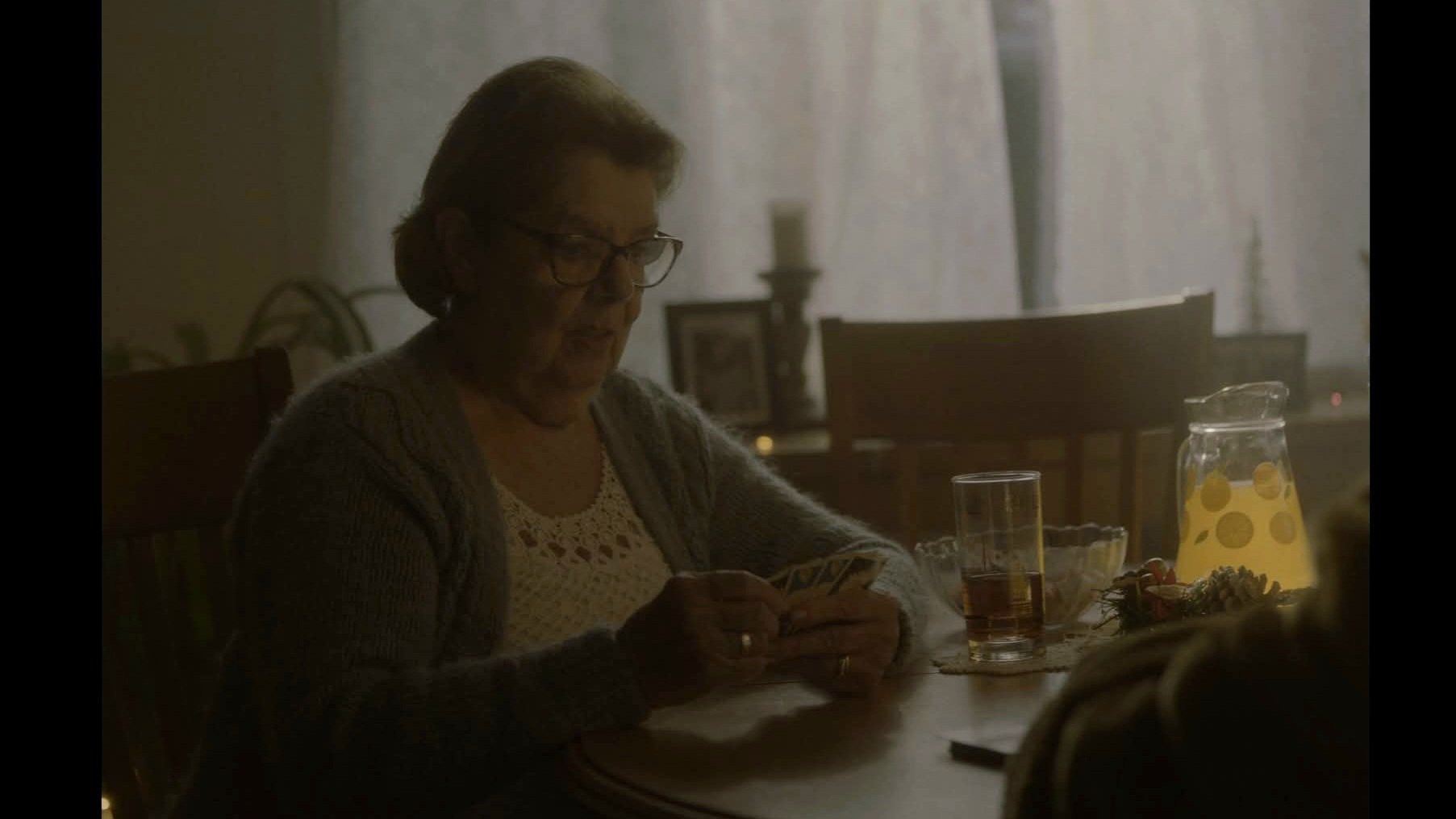 Une femme âgée assise à une table, regardant son téléphone, avec un pichet de limonade et un verre de boisson sur la table dans une pièce sombre et obscurcie.