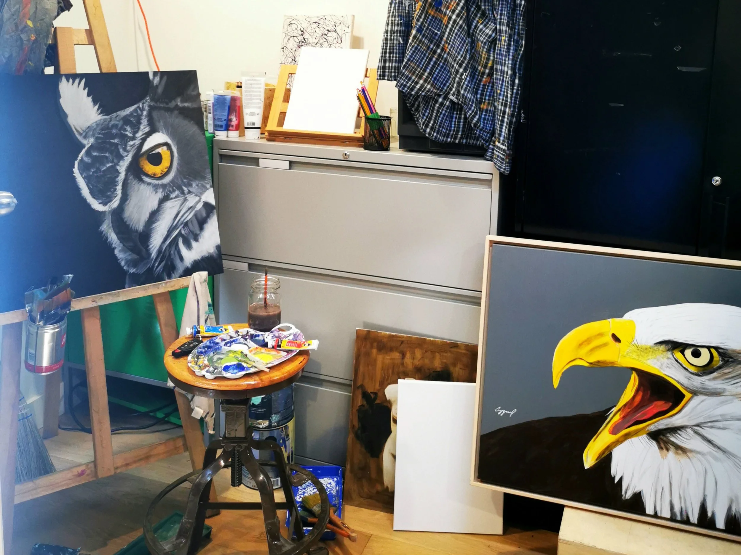 Studio d'art avec des œuvres en cours, y compris un portrait de hibou avec des yeux jaunes, des pinceaux, des tubes de peinture, une table avec de la peinture et des outils artistiques, et une toile blanche, créant un espace créatif.