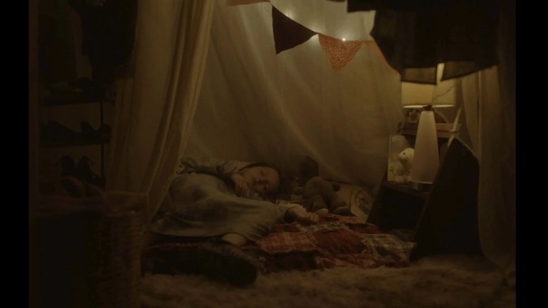 Une fille dort dans un lit avec des peluches dans une pièce chaleureuse et cosy, éclairée tamement avec une lampe de chevet, dans un environnement décoré avec des guirlandes lumineuses et des draps à carreaux.