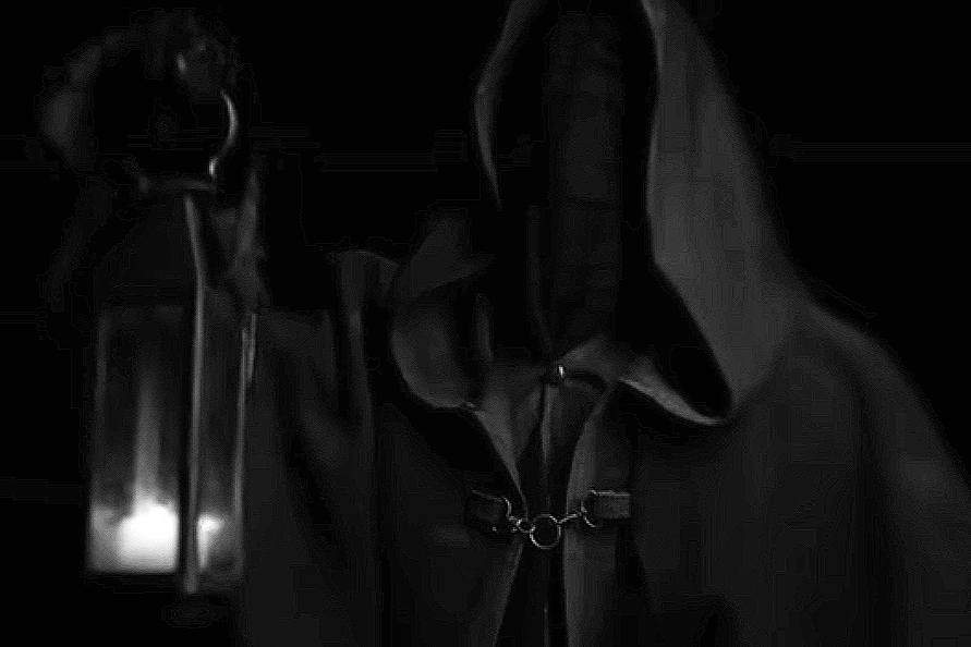 Deux personnes portant des t-shirts à capuche noirs, l'une avec la capuche sur la tête et l'autre sans. L'un porte une chaîne autour du cou. L'image est sombre, avec peu de détails visibles.
