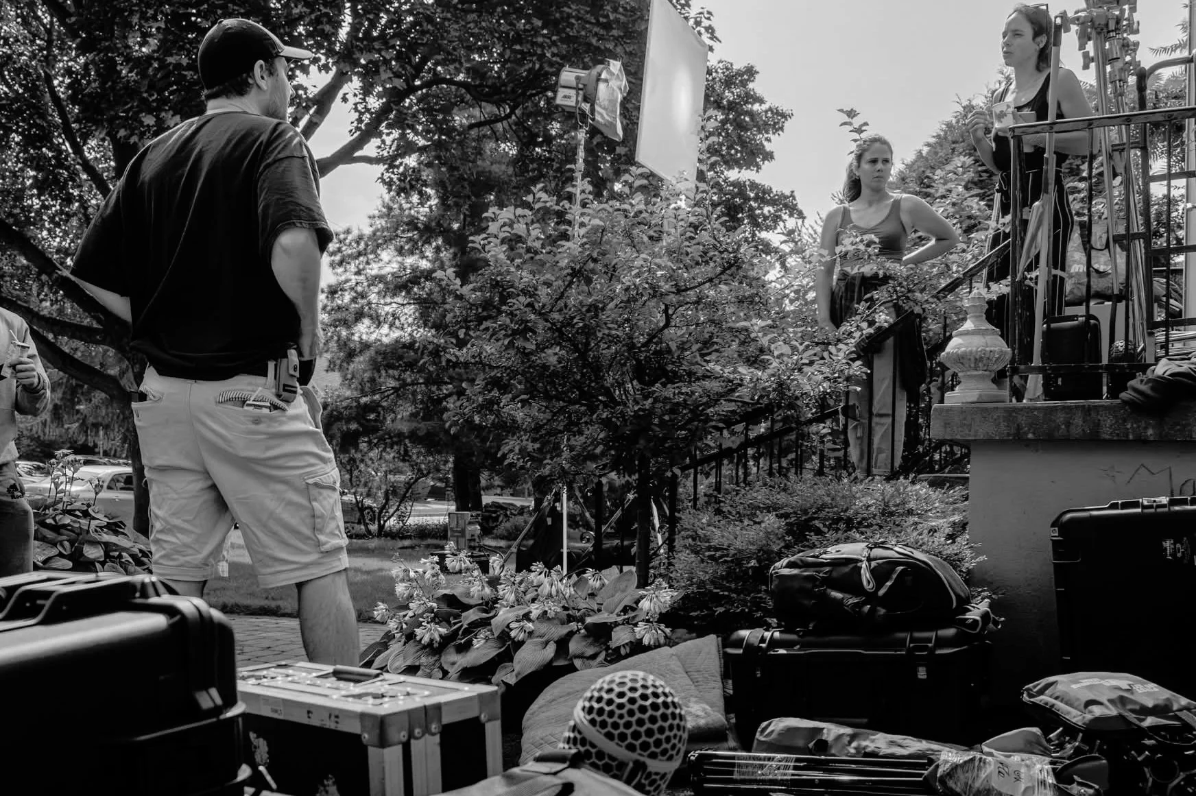Une scène de tournage en plein air avec une équipe technique préparant un tournage devant un décor de jardin. Deux femmes sont présentes : une debout en haut d'un ensemble de marches, tenant une tasse, l'autre devant, regardant pensivement. Divers éq