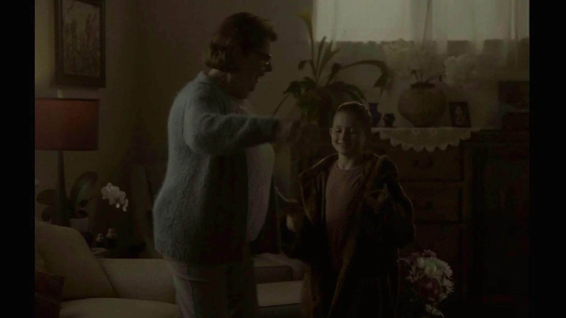 Une femme âgée et une jeune fille, toutes deux souriantes, dans un salon lumineux, échangeant un objet ou une poignée de main, avec des plantes et des décorations en arrière-plan.