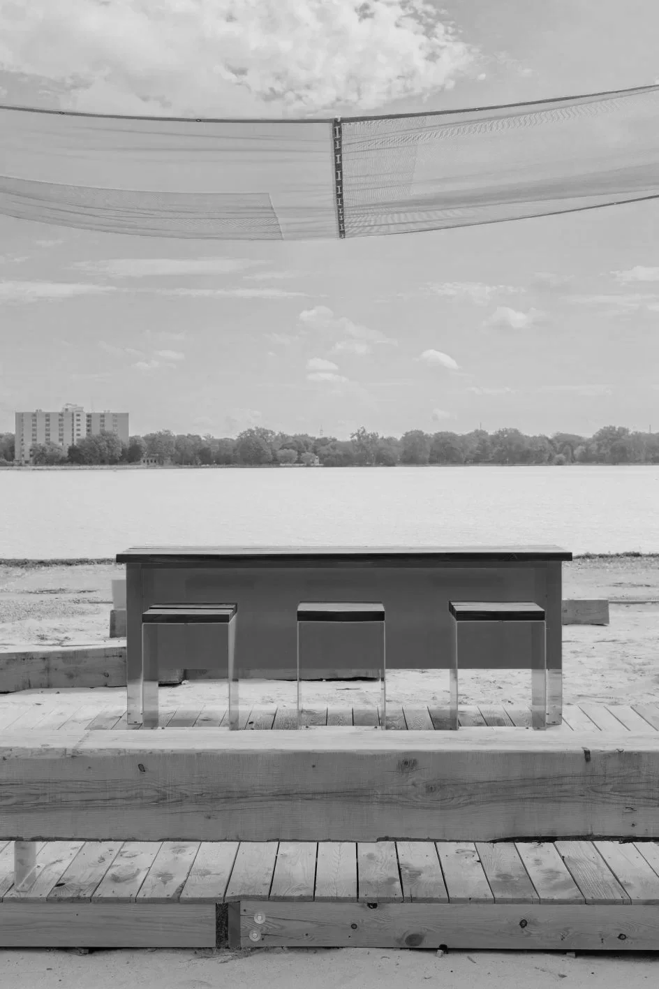 Un mobilier de table et de chaises en extérieur, près d'un lac avec des bâtiments en arrière-plan, sous un ciel partiellement nuageux.