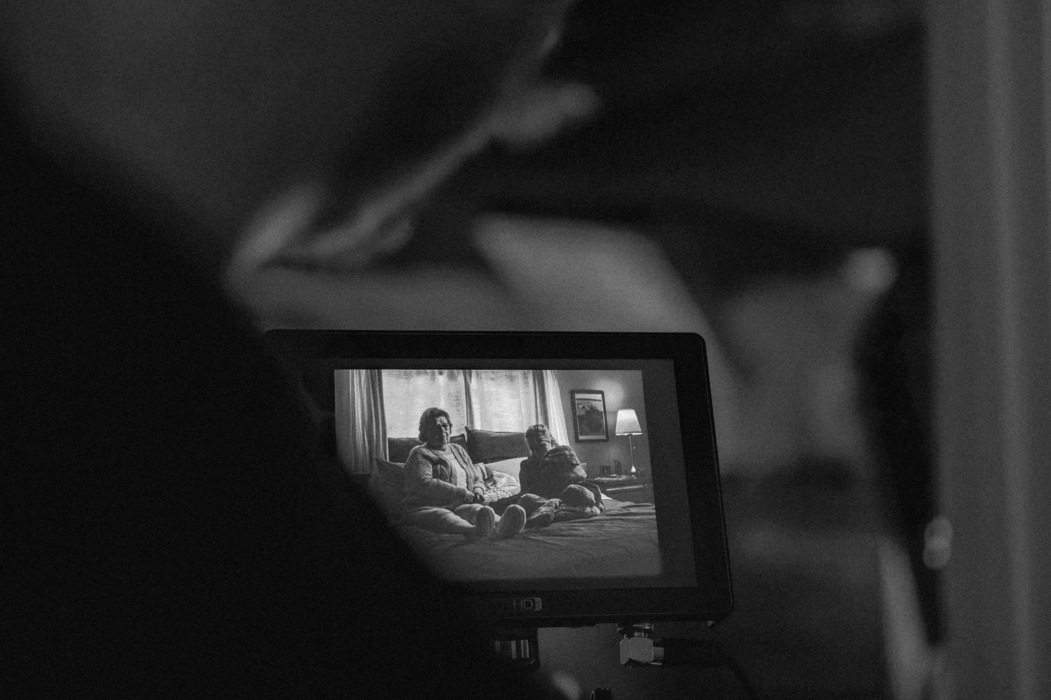 Une image en noir et blanc où l'on voit une personne prenant une photo d'un écran d'ordinateur. Sur l'écran, une scène de famille avec plusieurs personnes assises dans un salon en train de regarder la télévision.