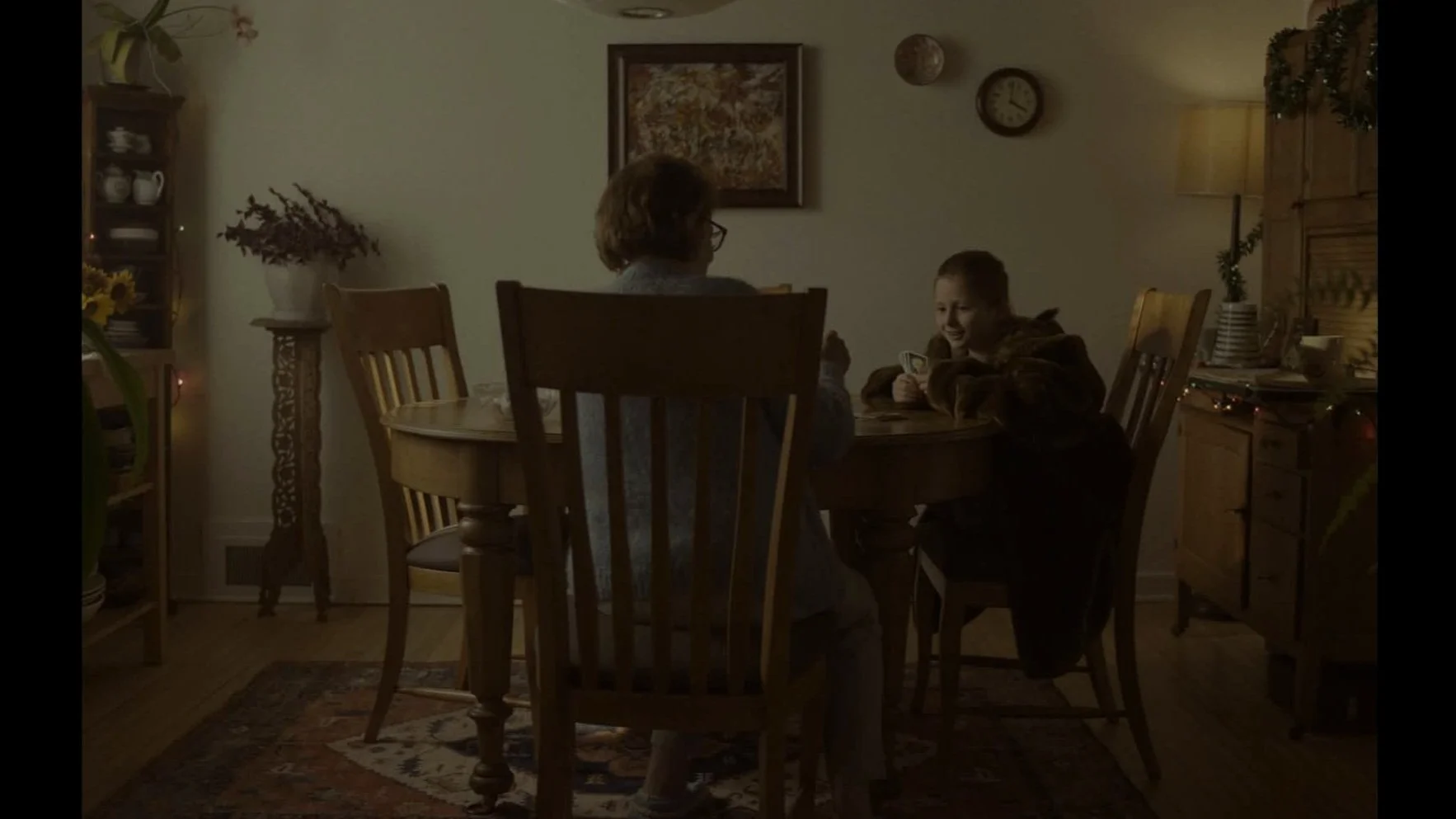 Une scène intime dans une salle à manger tamisée, deux personnes souriantes assises autour d'une table en bois, jouant à un jeu de cartes. La pièce est décorée avec des fleurs, des étagères, une horloge, et des guirlandes lumineuses, créant une ambia