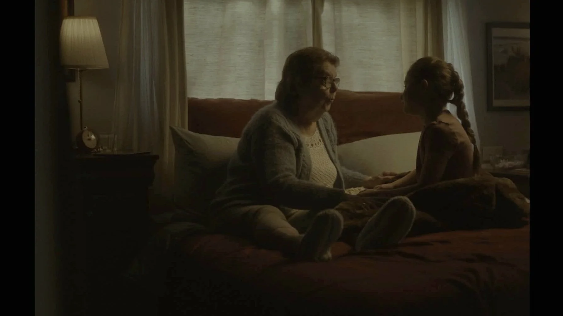 Une grand-mère et une petite fille assises sur un lit dans une chambre tamisée, tenant la main et se regardant dans les yeux.