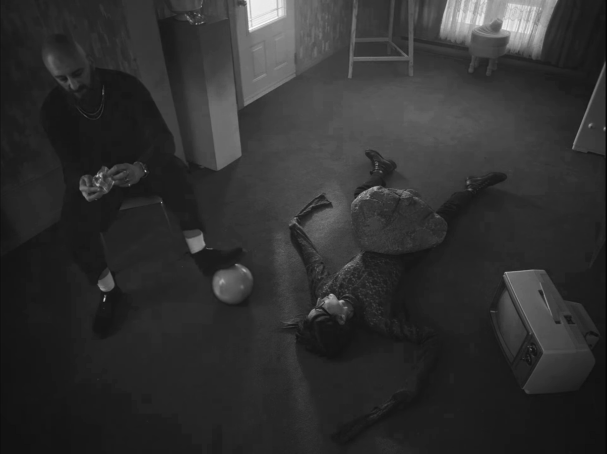 Une scène sombre où une femme est allongée sur le sol avec une roche sur le ventre et un homme assis à côté, tenant un objet dans ses mains. La pièce semble être une salle à vivre avec des meubles et une télévision.