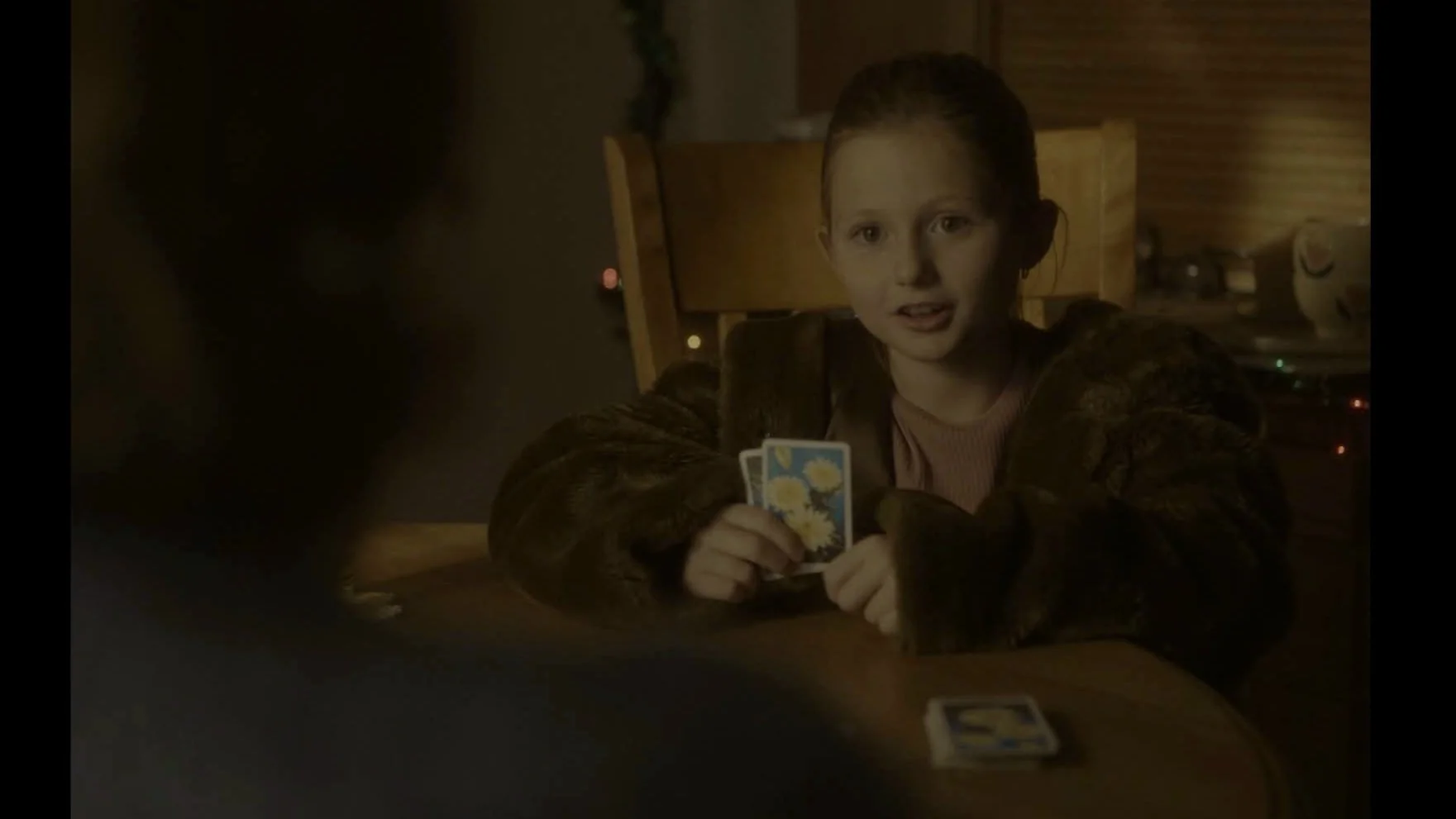 Une jeune fille aux cheveux roux, portant un manteau sombre, joue aux cartes à une table dans une pièce avec un éclairage tamisé.