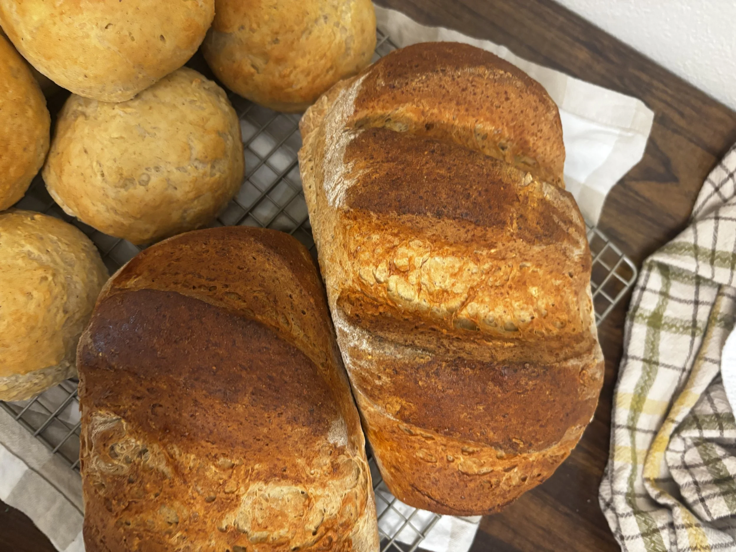 Close-up of loaves of bread, some with a dark crust and some with a lighter, yellowish crust, resting on a cooling rack and a checkered cloth.