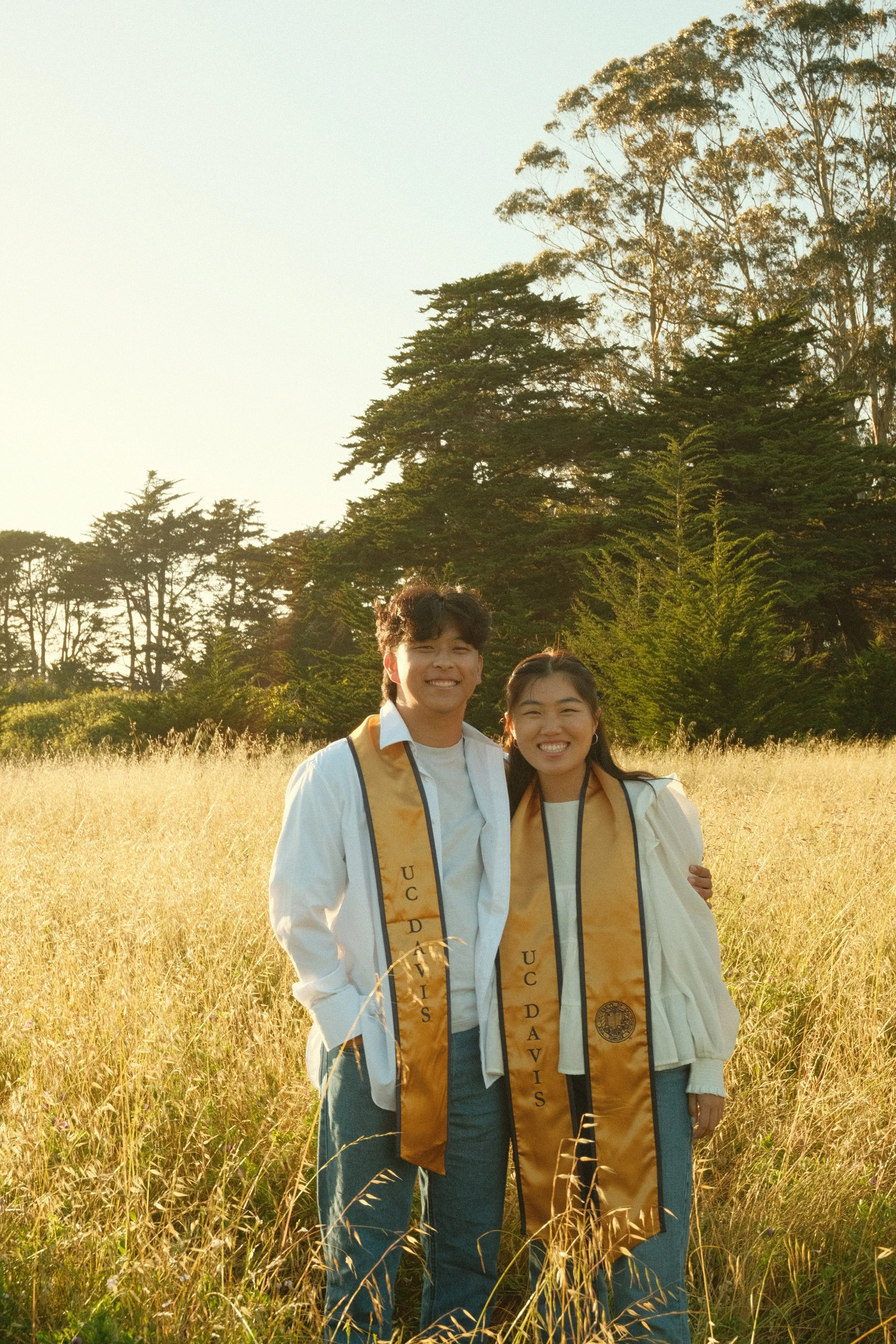 Two young adults in graduation caps and gowns standing in a field of tall grass with trees in the background during sunset, smiling at the camera.