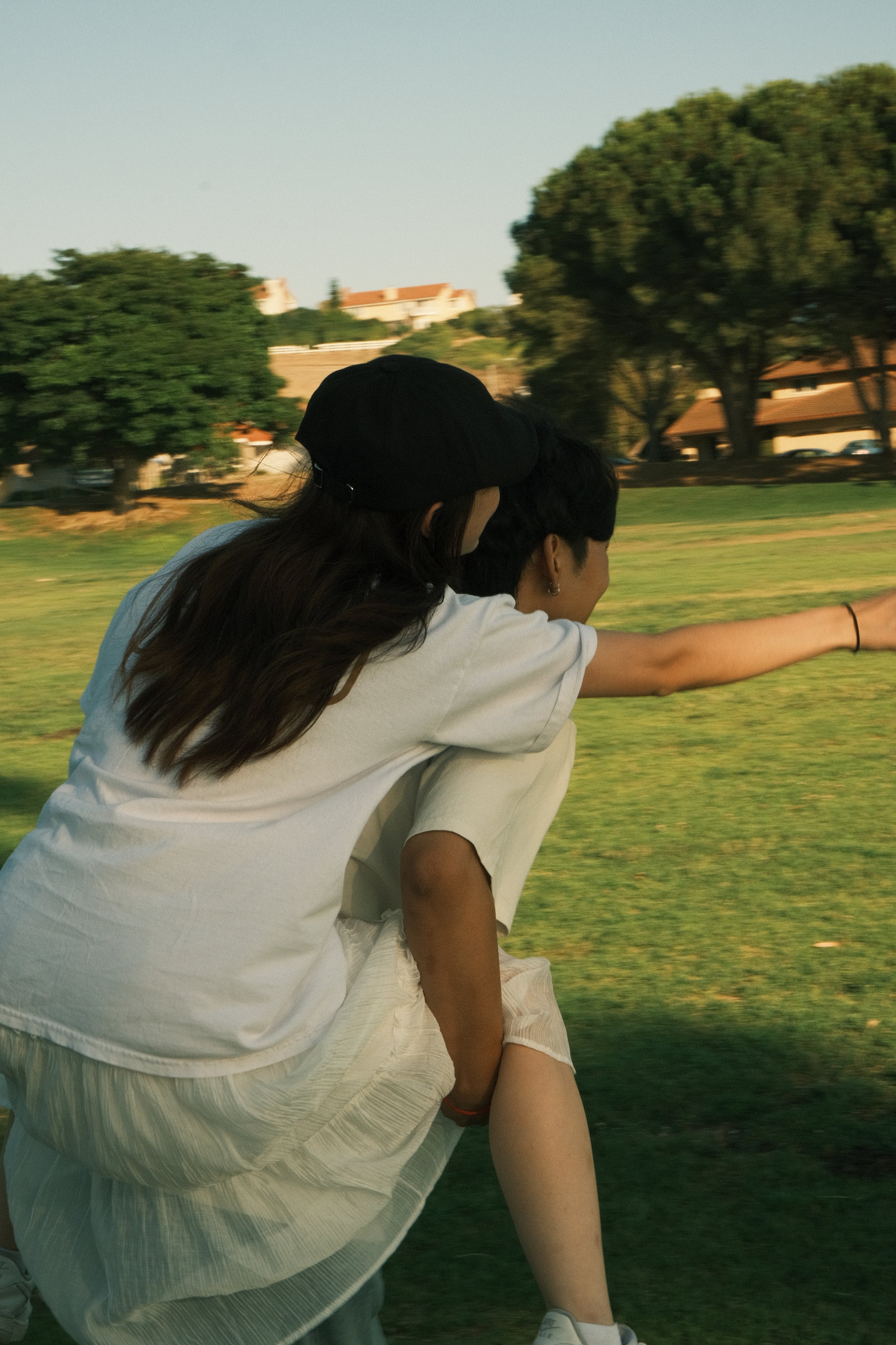Two young women riding a bike in a park with green grass, trees, and buildings in the background during daytime.