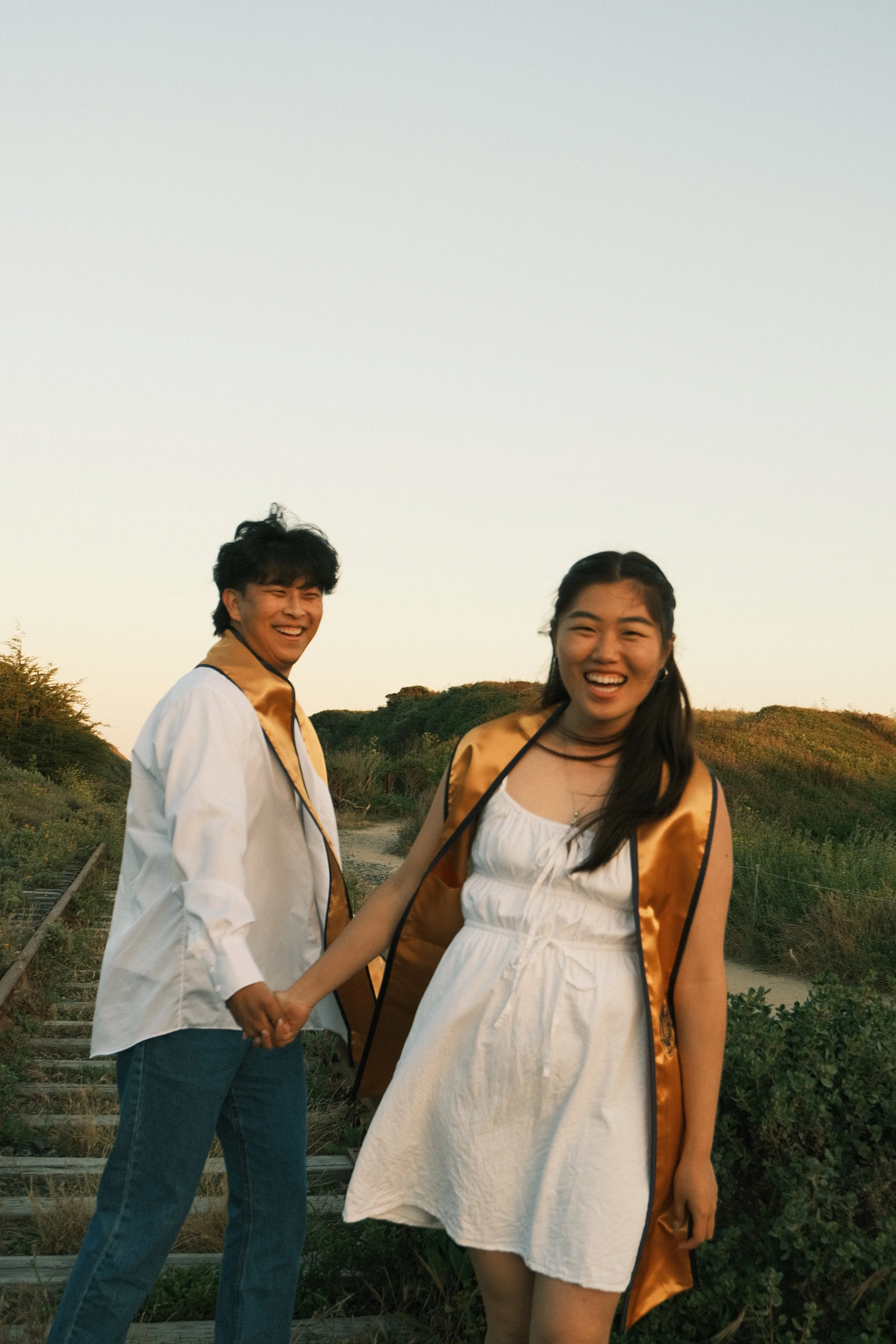 A young man and woman holding hands outdoors during sunset, smiling and looking at the camera, with a scenic hill and greenery in the background.