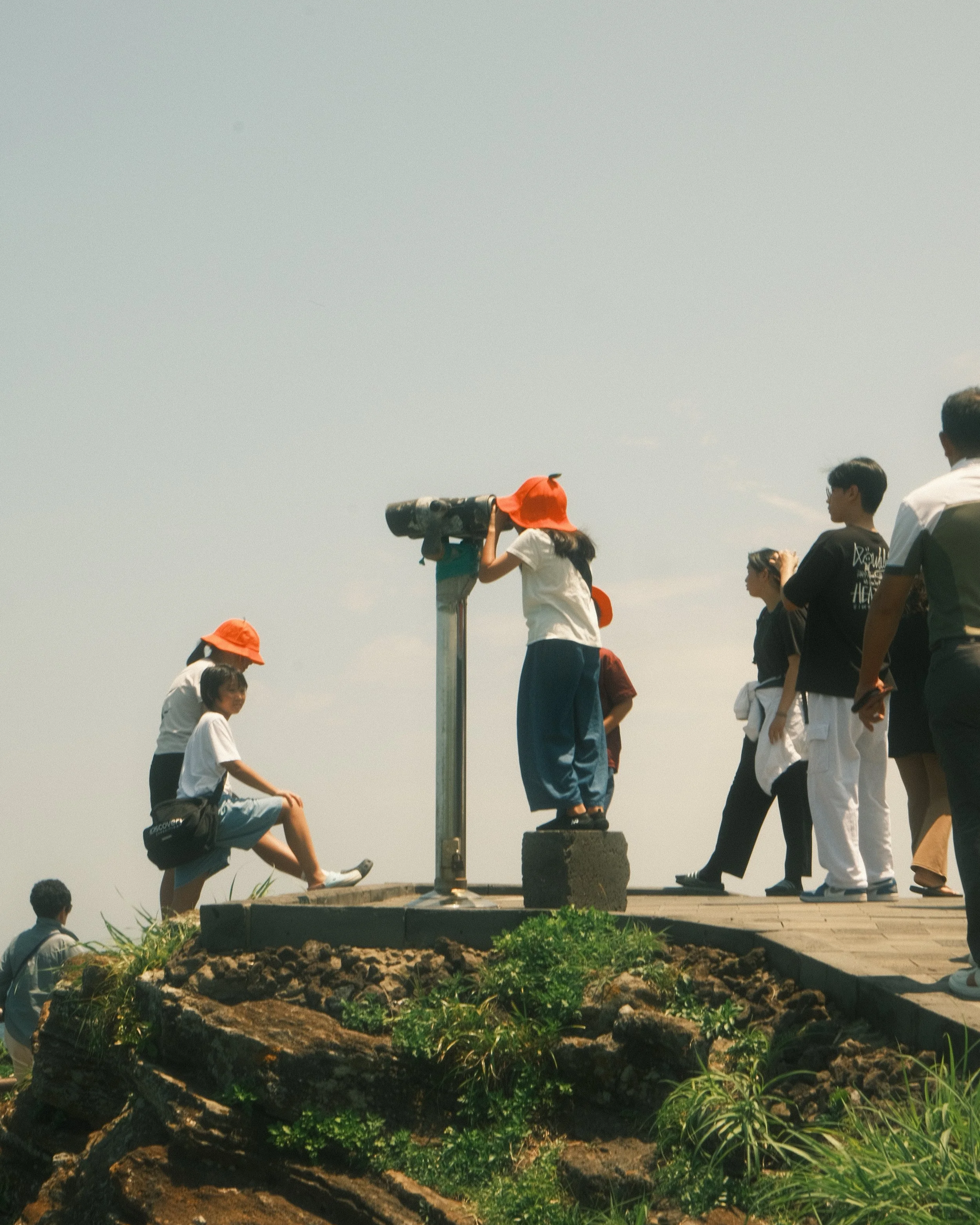People gathered outdoors during daytime, with some standing and others sitting, observing or engaging in an activity. A girl is using a telescope mounted on a pole, with a large orange hat, against a clear sky.