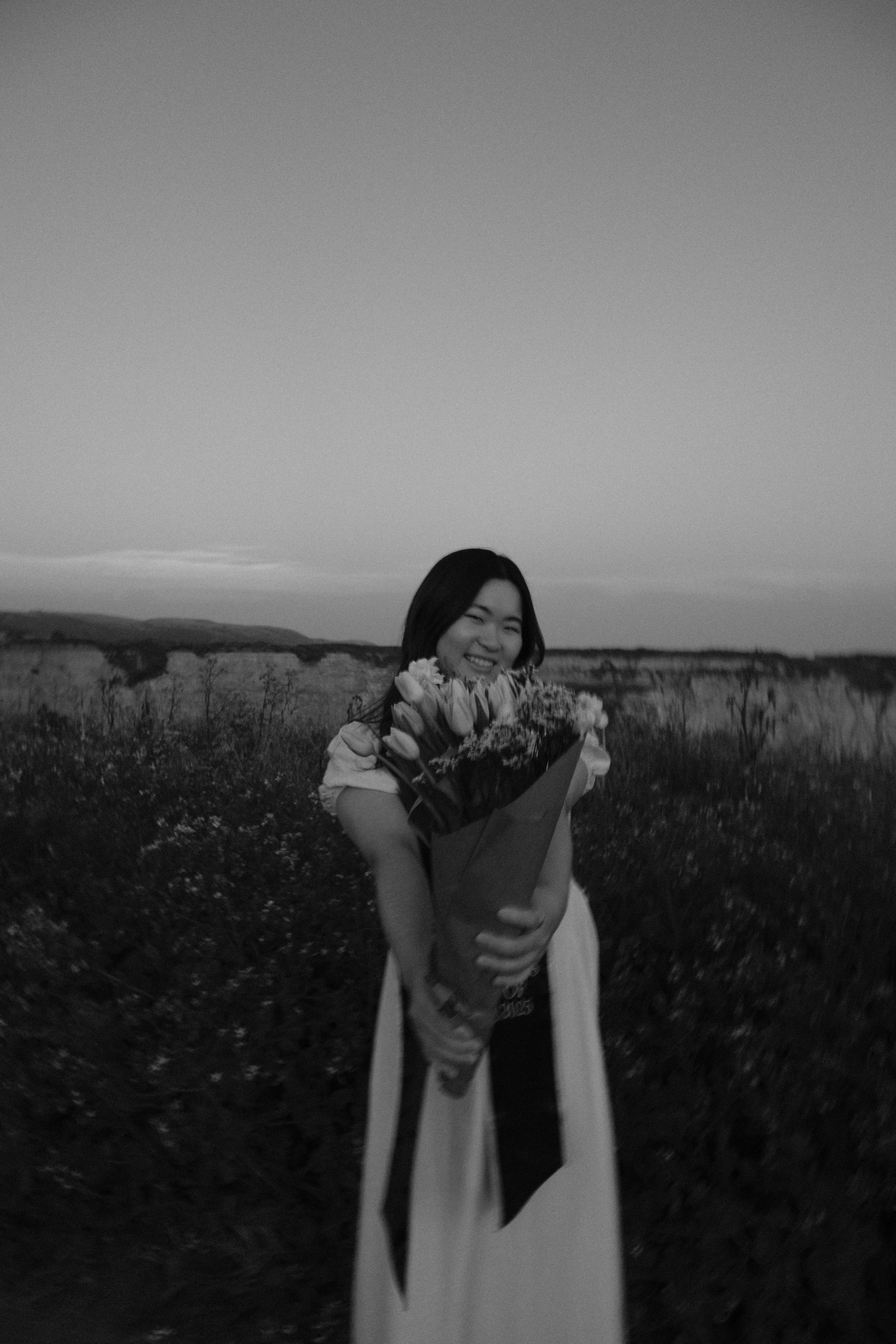 A woman smiling and holding a large bouquet of flowers outdoors during sunset or dusk, with a landscape of fields and hills in the background.
