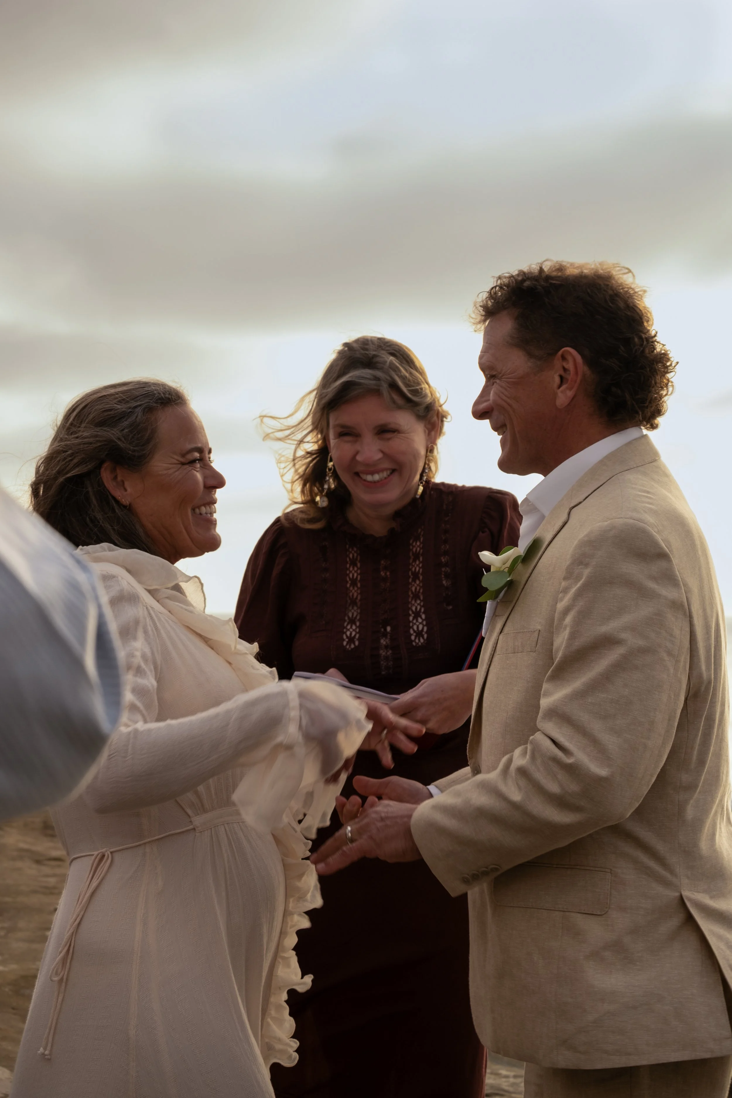 A couple is exchanging vows during a beach wedding ceremony, holding hands and smiling at each other, with a woman officiant standing between them and looking at the couple, all under an overcast sky.