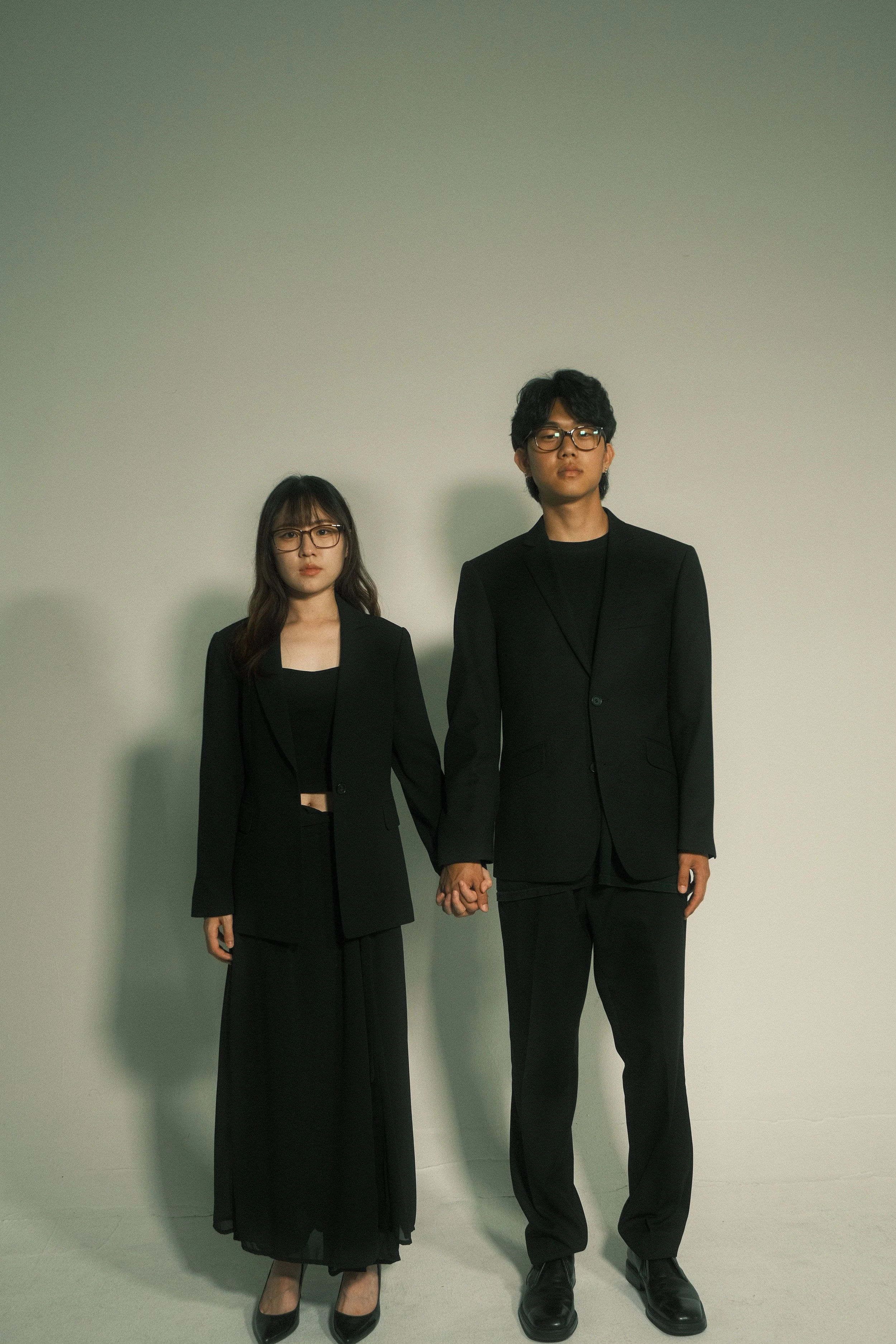A man and woman standing against a plain light green wall, holding hands. Both are dressed in black suits, with the woman wearing a crop top and long skirt, and the man in a blazer and dress shoes. They are wearing glasses and have serious expressions.