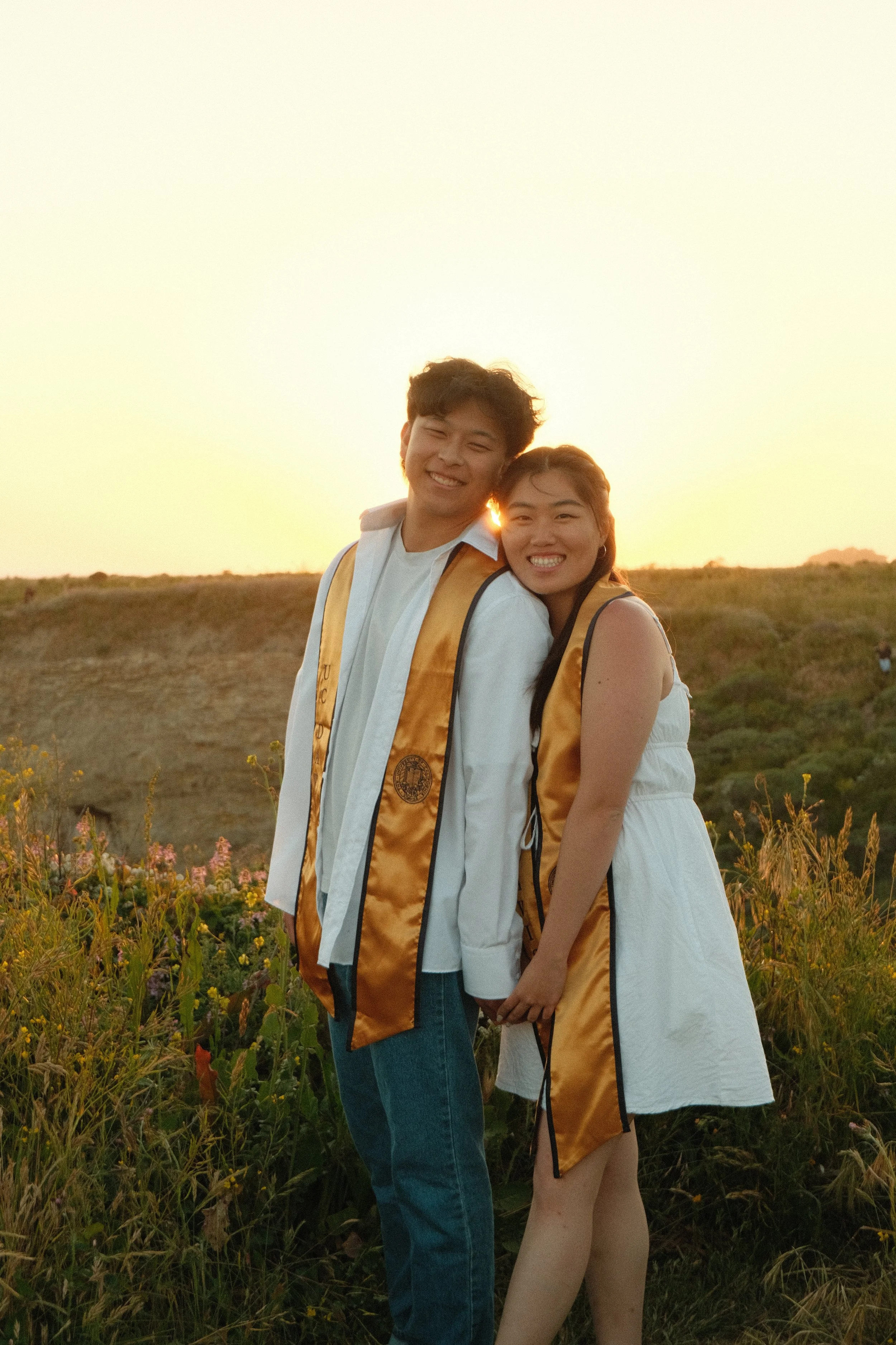 A young man and woman are standing in a field with wildflowers at sunset, both wearing graduation stoles and smiling at the camera.