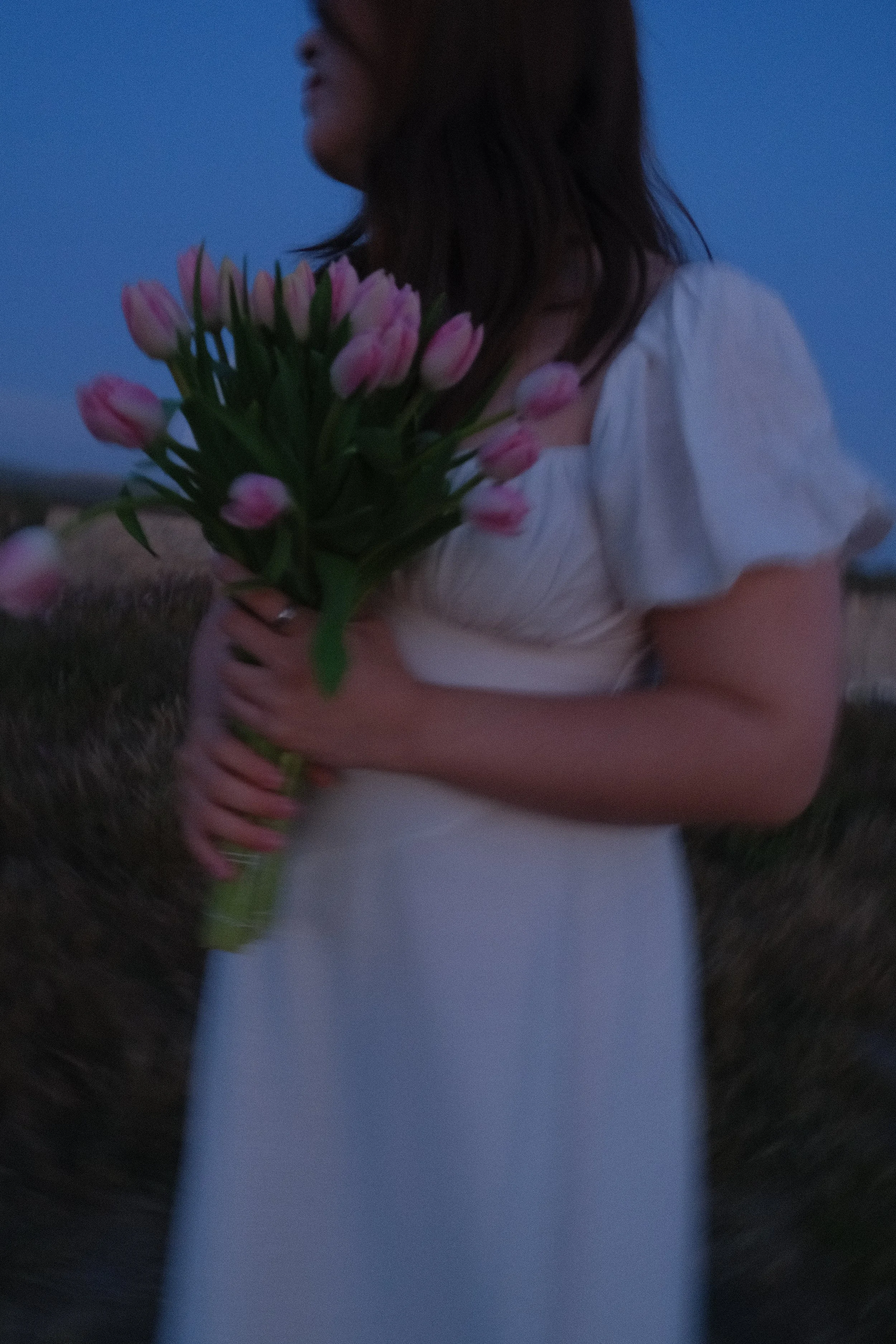 A woman with brown hair holding a bouquet of pink tulips, wearing a white top, standing outdoors during dusk or early evening.