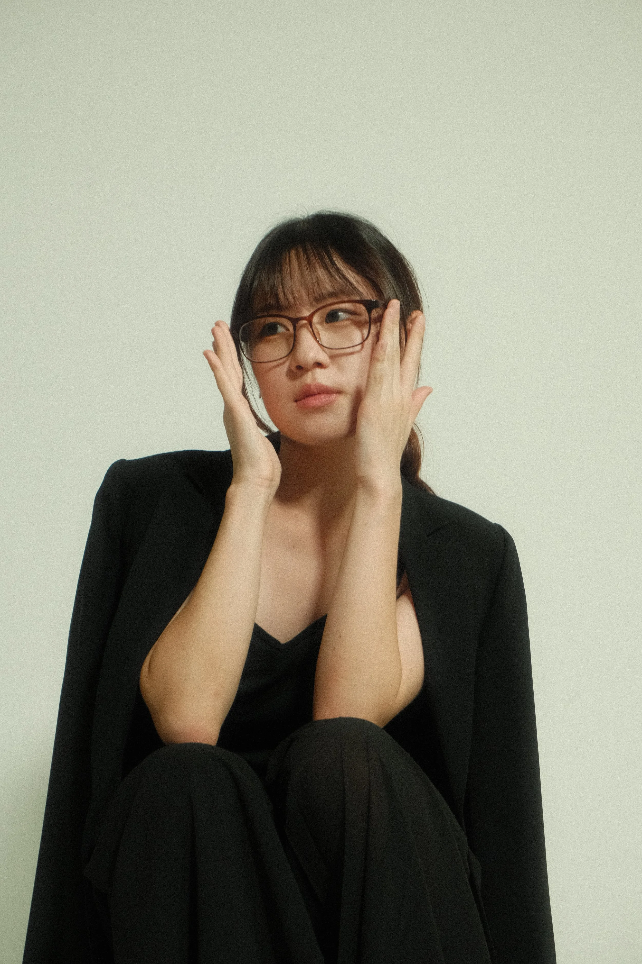 A woman with dark hair, glasses, and a black blazer is sitting against a white background, touching her face with both hands and looking upwards.