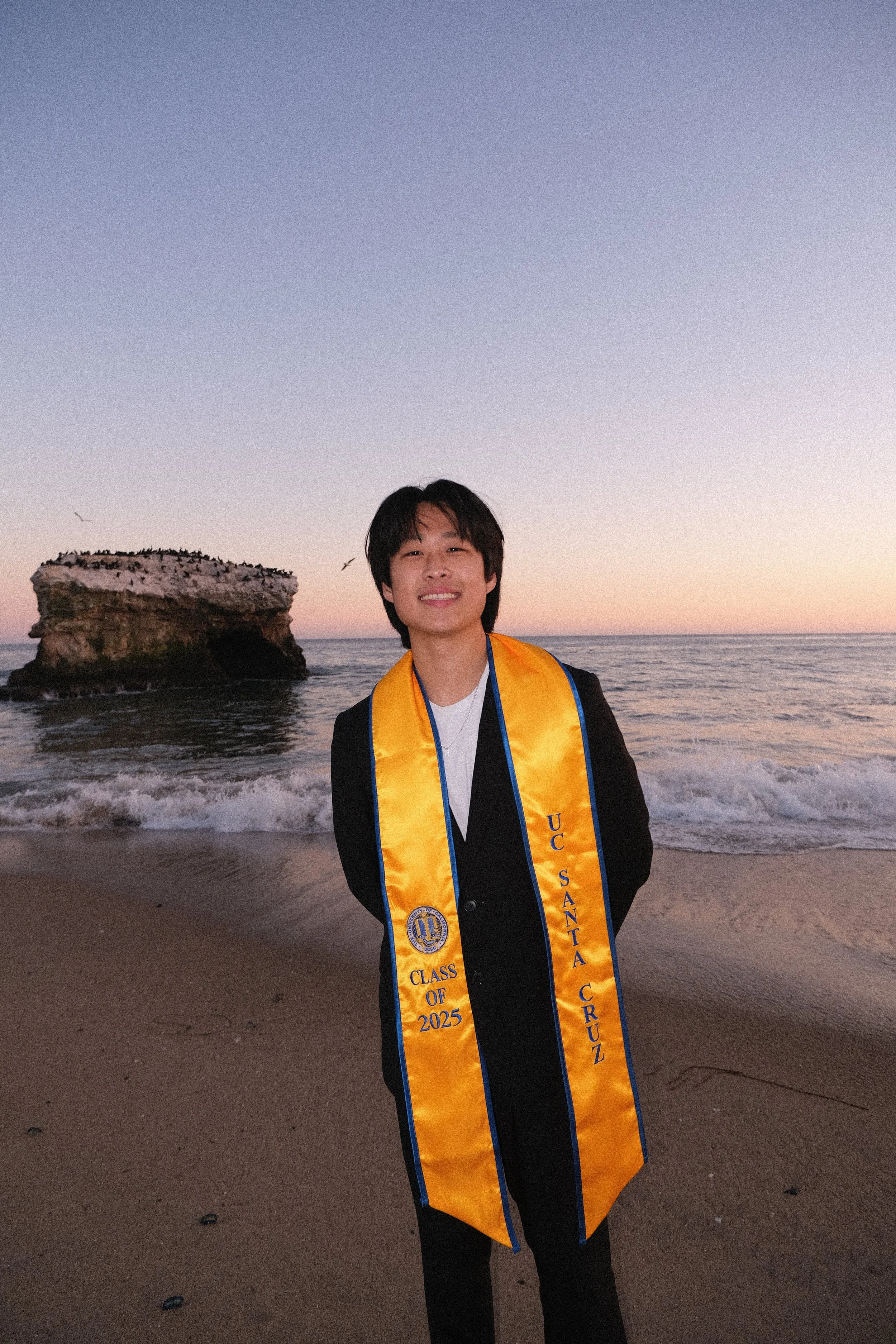 A young man smiling on the beach at sunset, wearing a graduation stole that reads 'Class of 2025 Santa Cruz' over a suit.