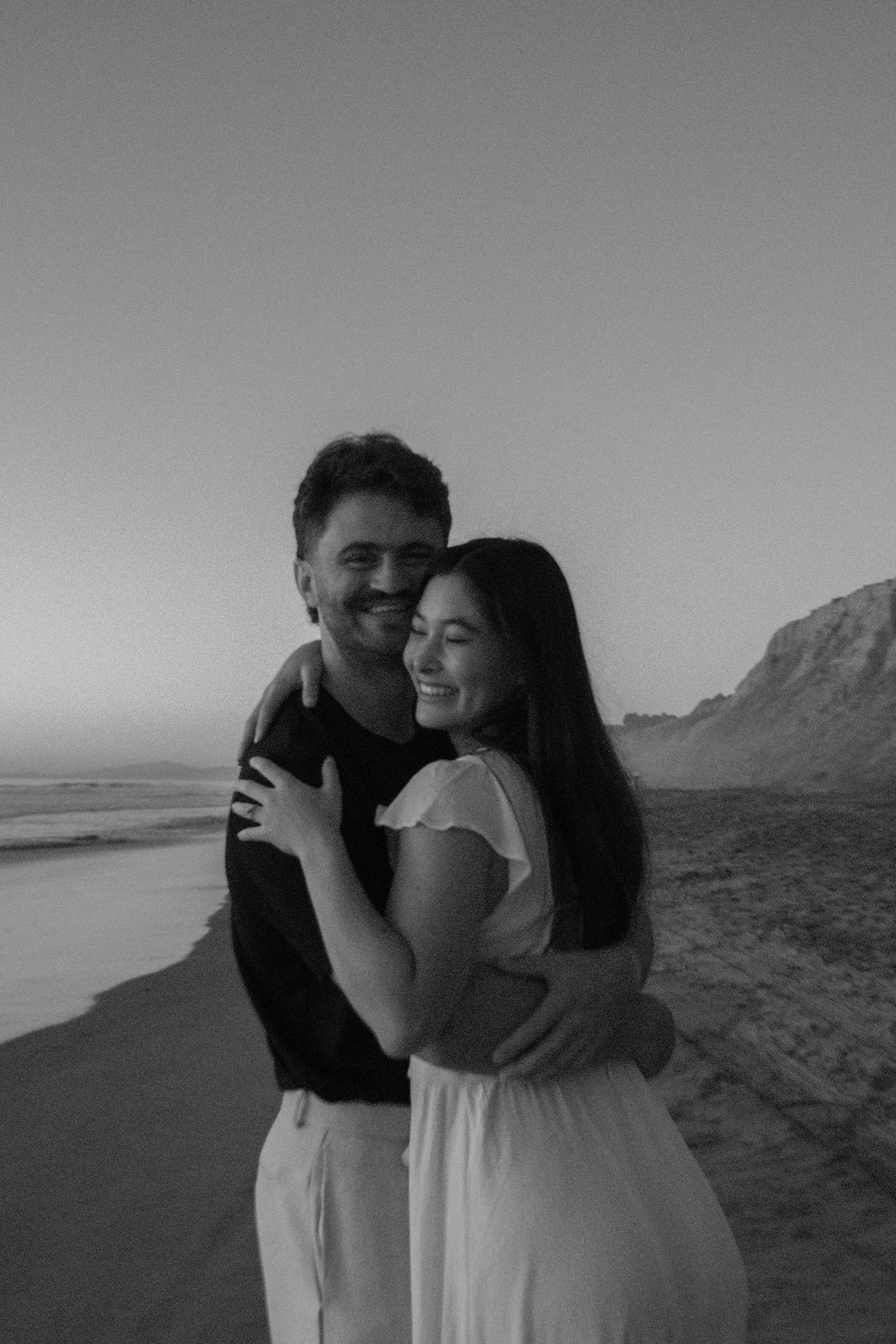 A black and white photo of a smiling young man and woman hugging on a beach with ocean waves and rocky formations in the background.