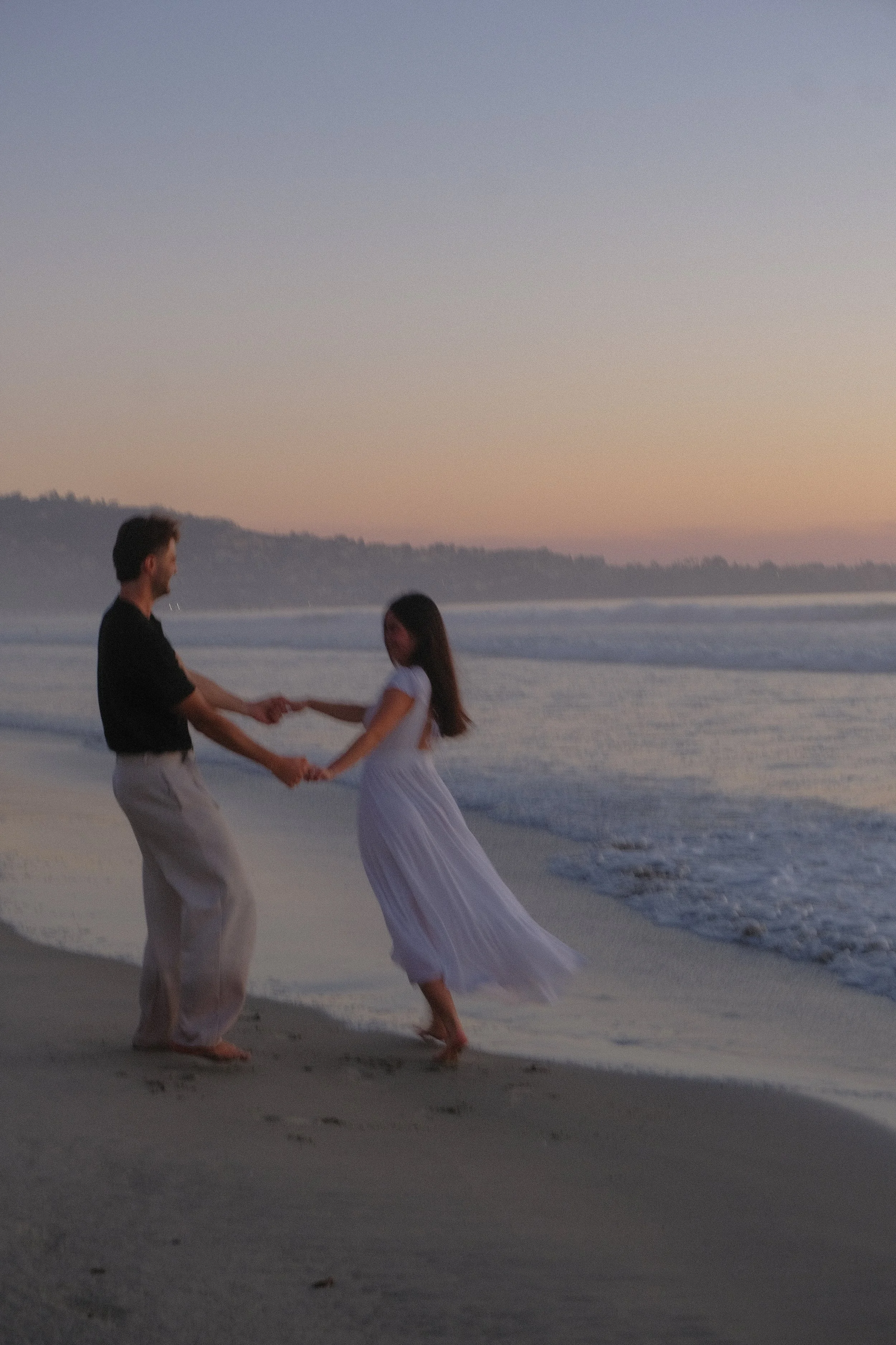 A couple holding hands and dancing on the beach at sunset, with waves in the background and a distant shoreline.