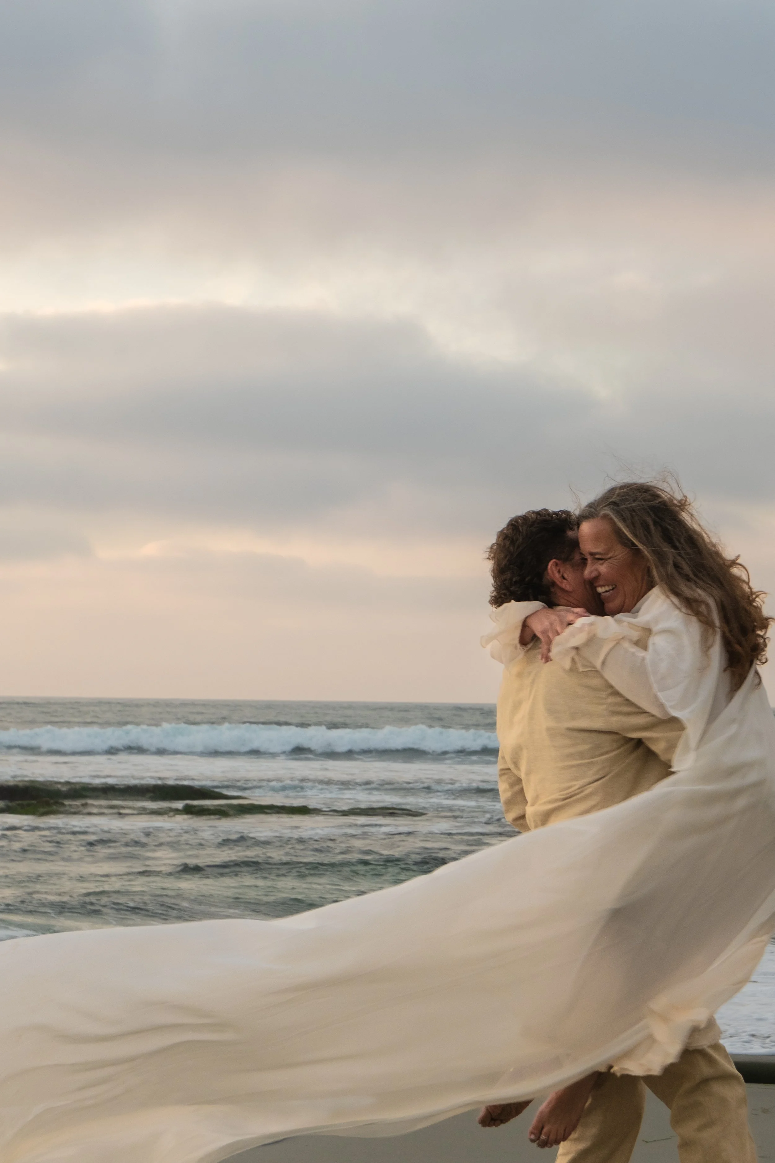 A couple dressed in light-colored clothes embrace and smile on a beach with waves and a cloudy sky in the background.