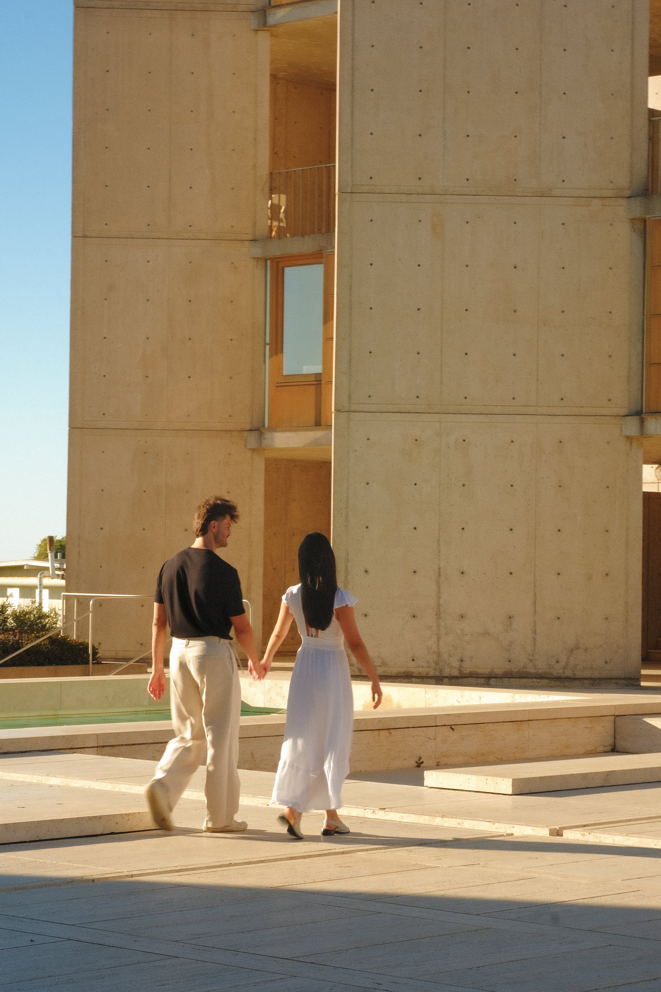 A couple is holding hands and walking outside near a modern concrete building with multiple windows and wooden accents. The man is wearing a black t-shirt and light-colored pants, and the woman is wearing a white dress.