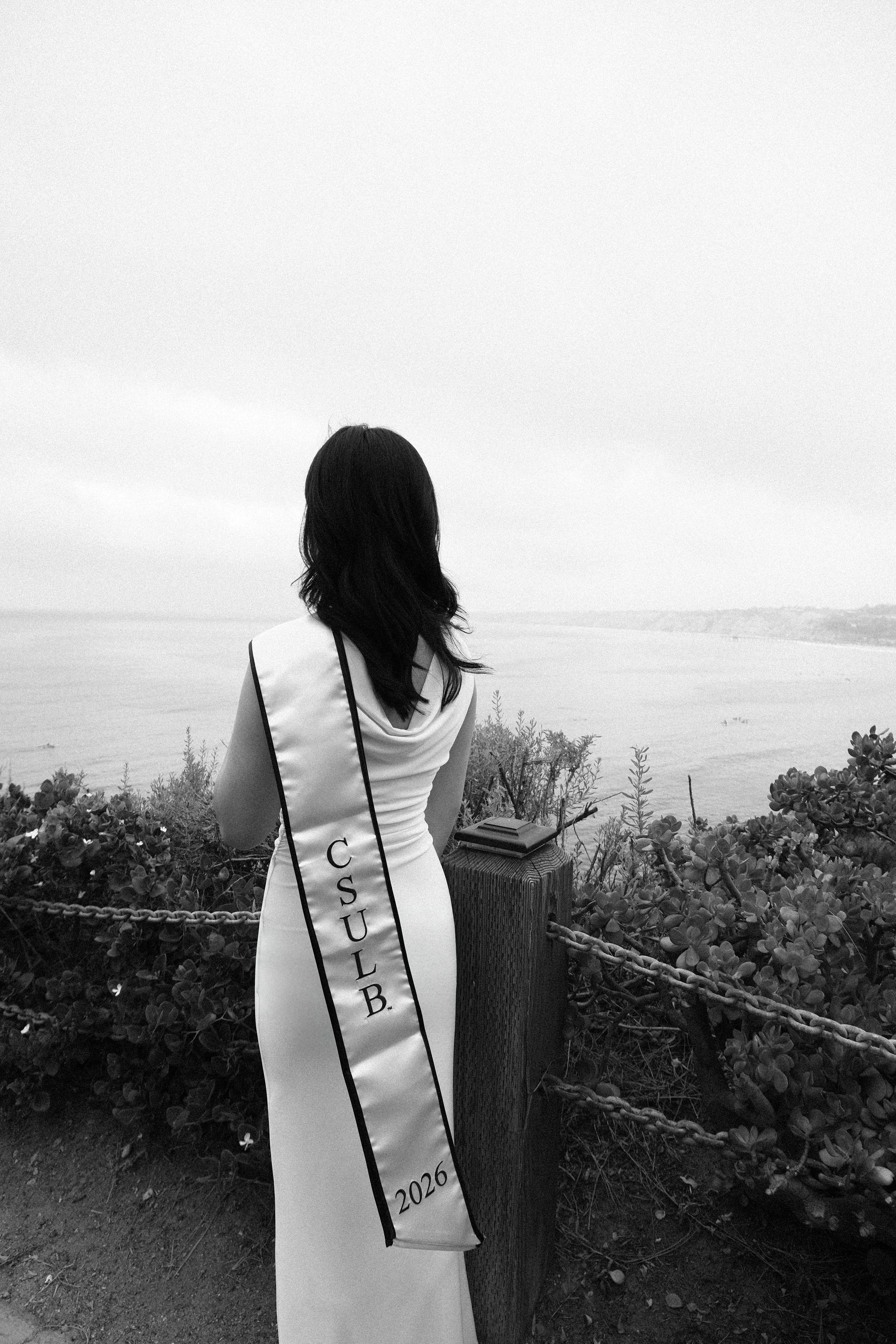 A person with shoulder-length hair wearing a sash that reads 'COSTUB' stands near a wooden post and a chain in front of shrubs and a body of water. The person's back is to the camera, facing the water and distant shoreline.