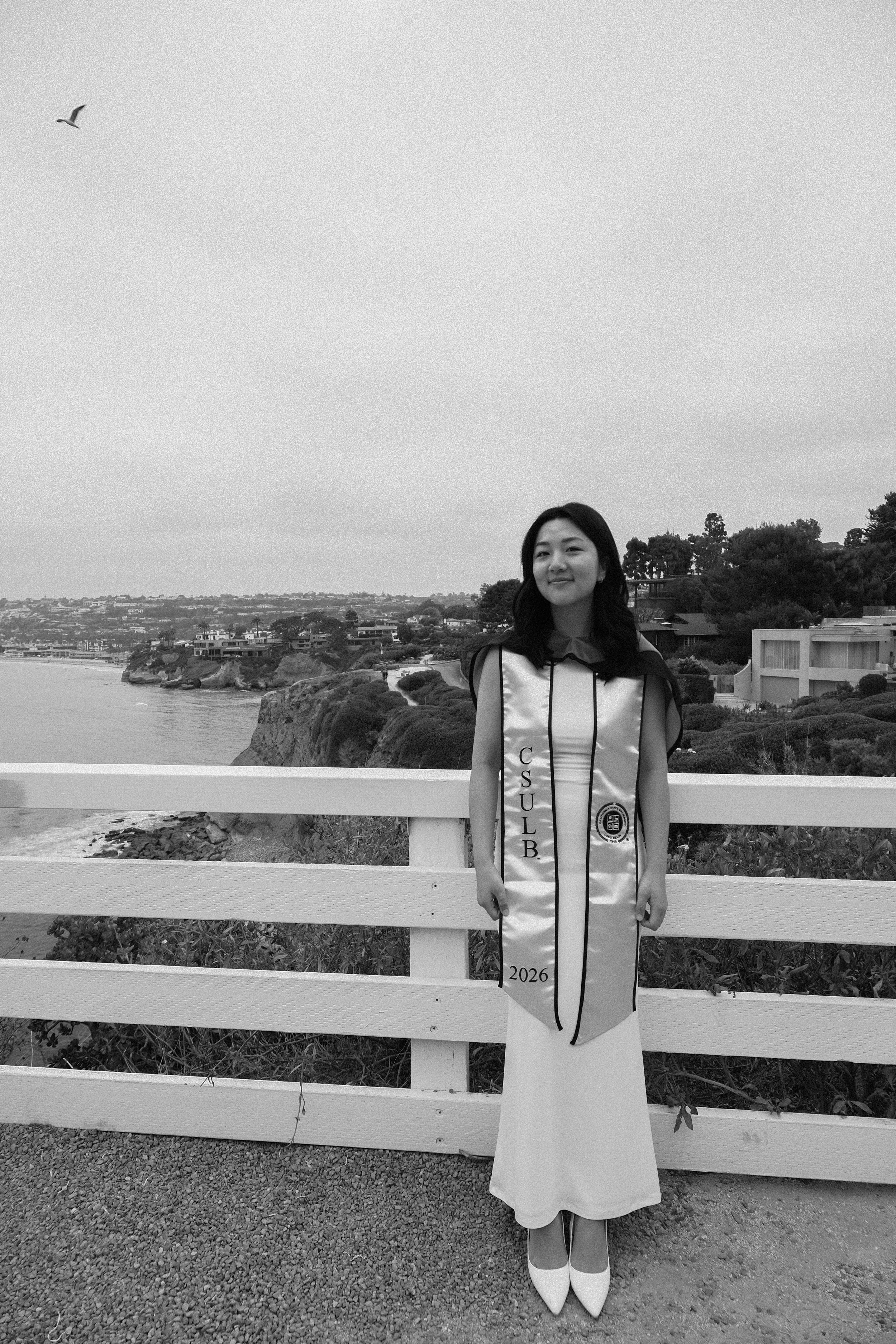 A woman dressed in a gown and sash that reads 'CSULB 2026' stands behind a white fence by the coast, with houses and trees in the background and a seagull flying overhead.