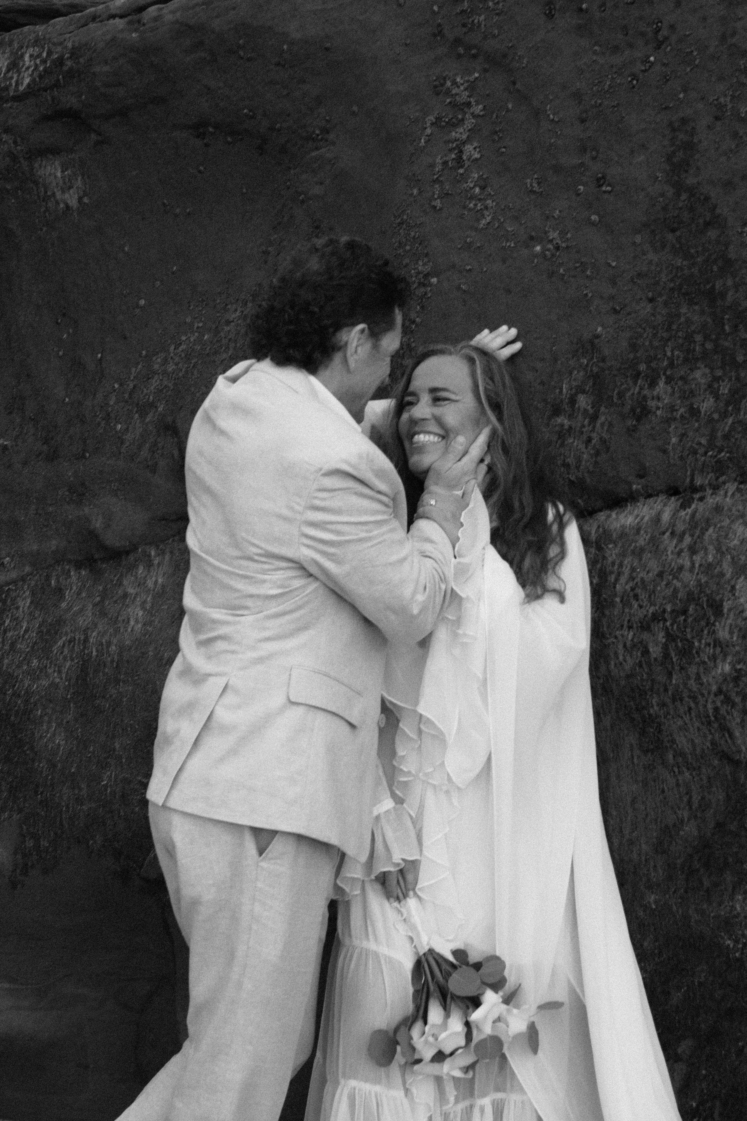 A man and woman are smiling and looking at each other, with the man touching the woman's face and the woman holding a bouquet of flowers, standing outdoors near rocks.