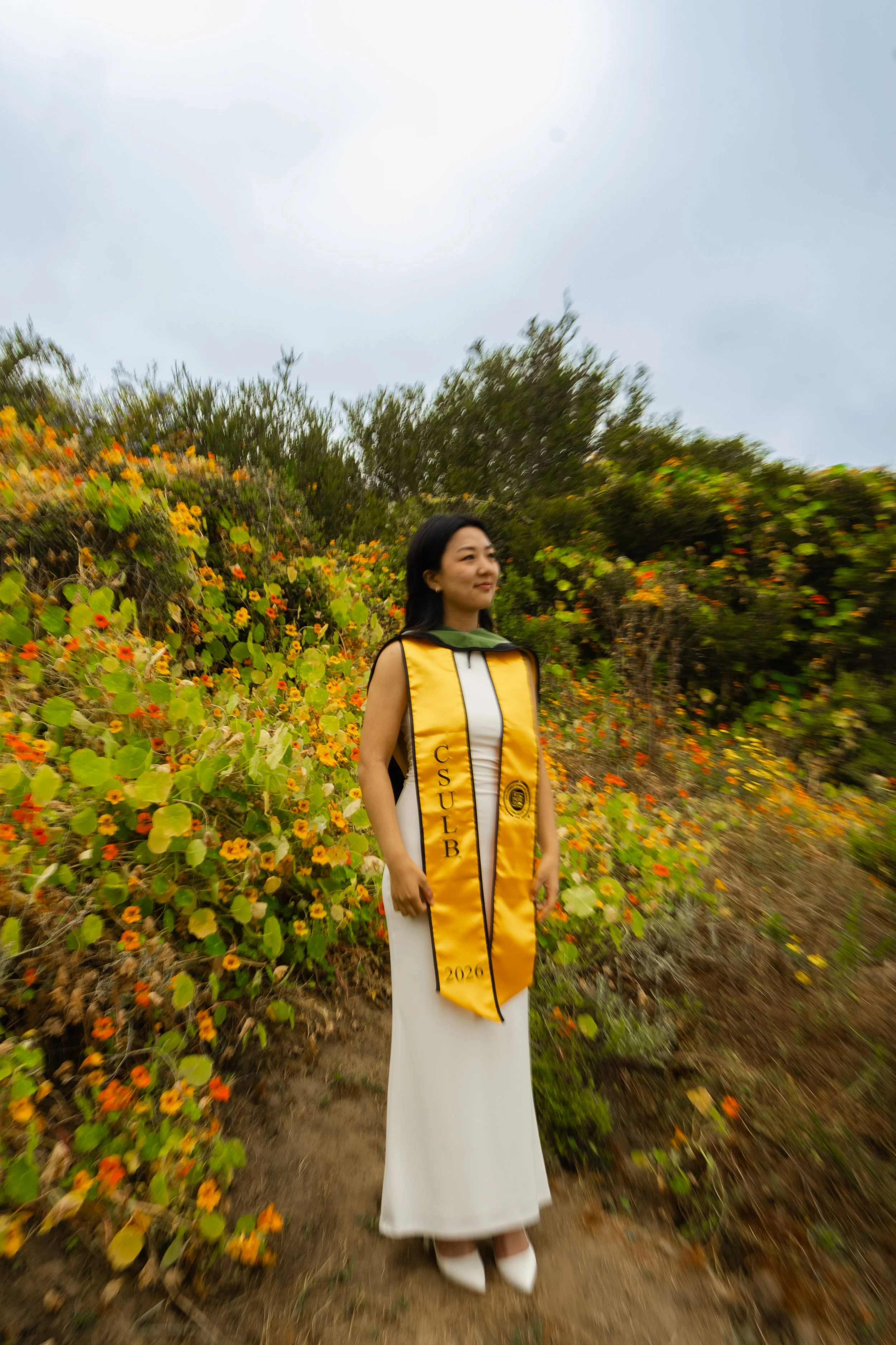 A woman in a white dress and a yellow sash stands outdoors on a dirt path surrounded by orange and yellow flowers and green bushes, under a cloudy sky.