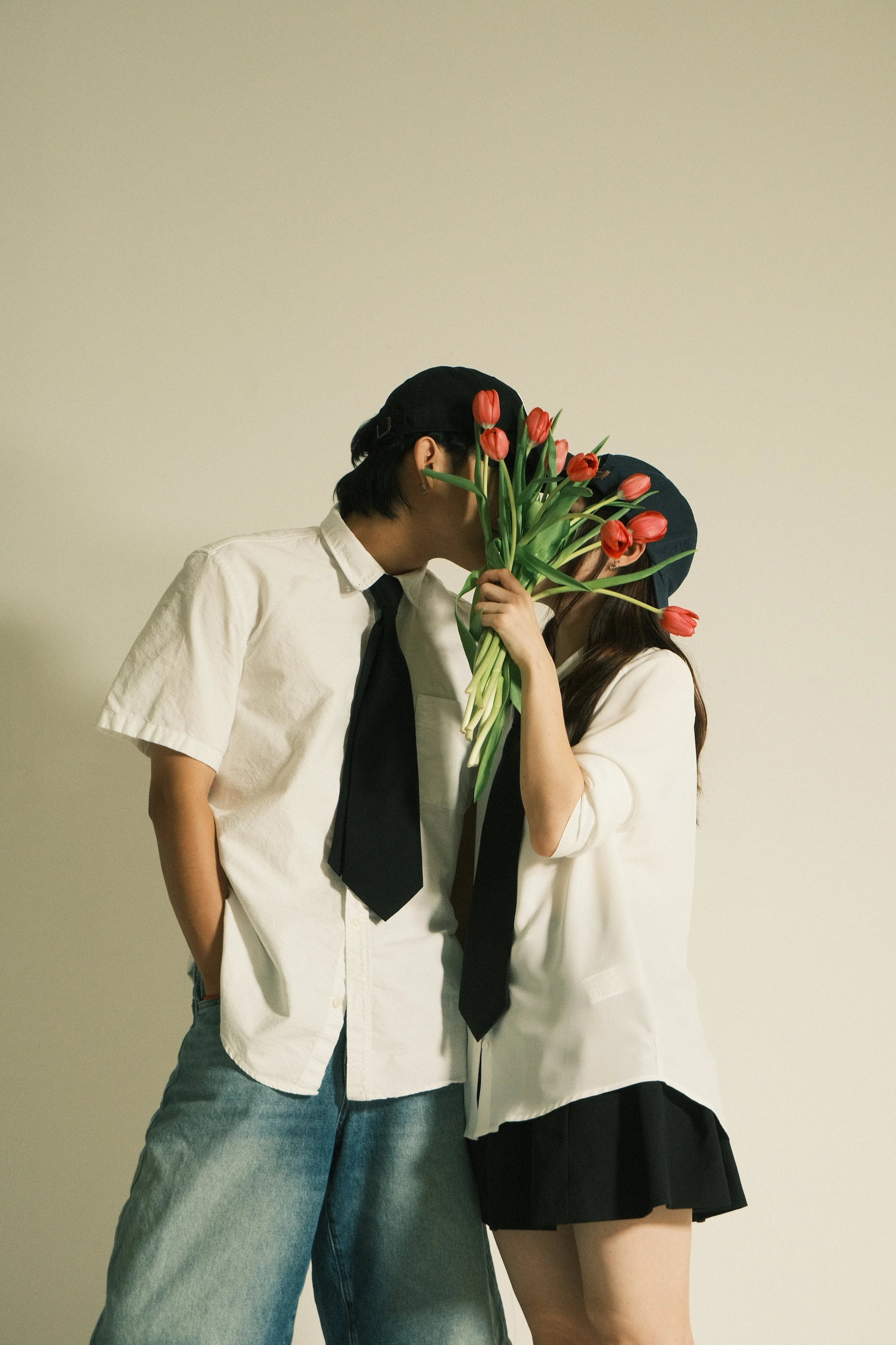 Two people dressed in white shirts and black ties, facing each other, holding a bouquet of red tulips between them, with one person covering part of their face with the flowers, against a plain light-colored background.