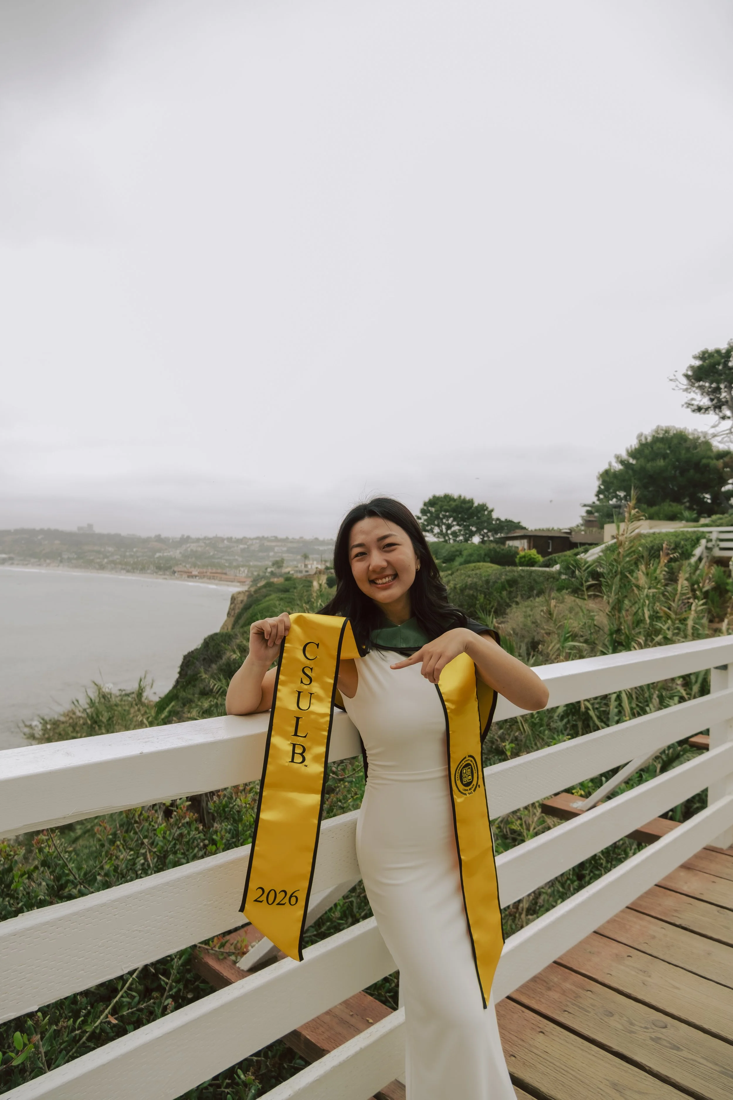 A smiling woman wearing a white dress and a graduation sash with the year 2026, standing beside a white wooden fence overlooking a coastal landscape with greenery, trees, and cloudy sky.