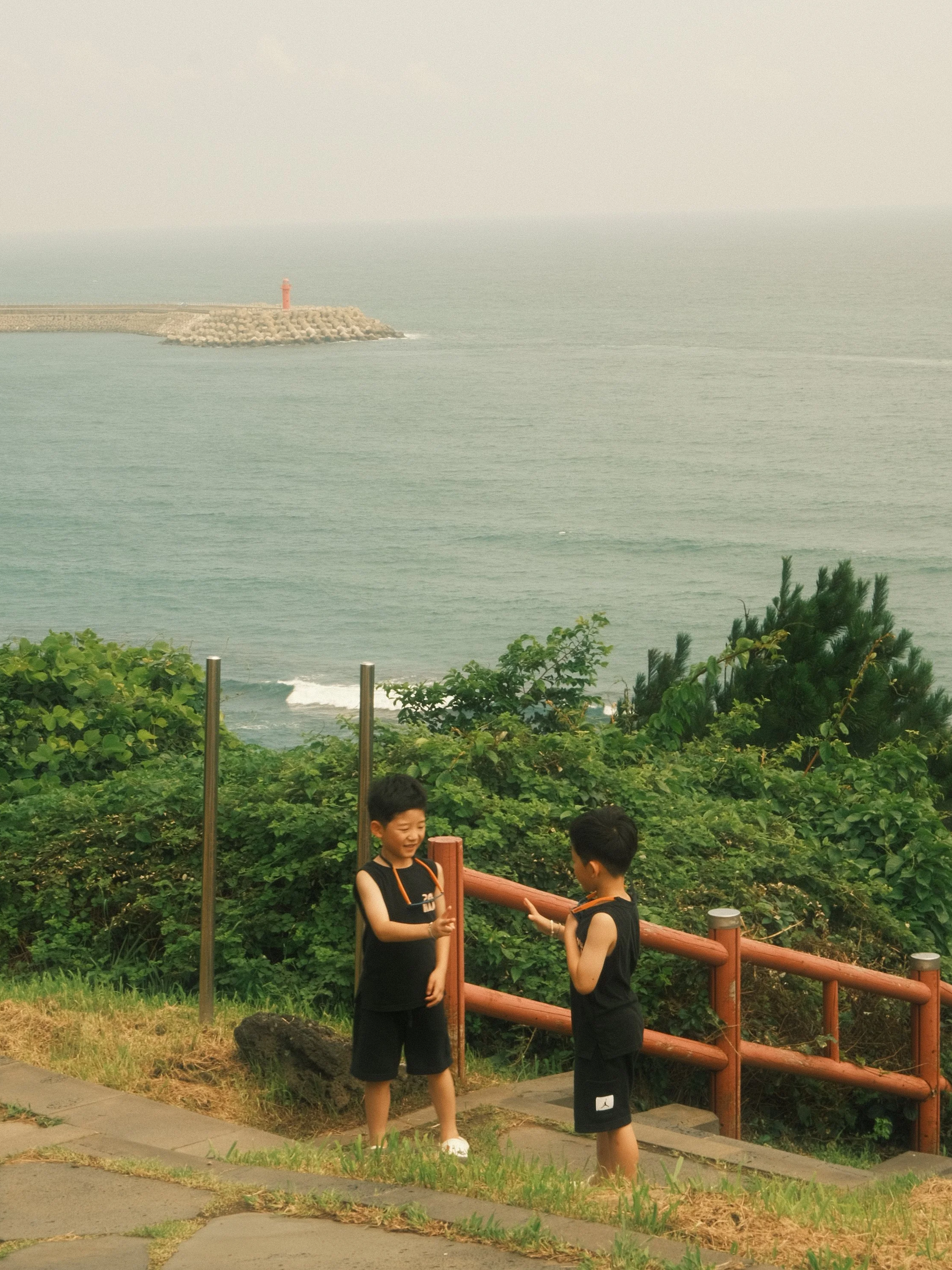 Two boys in black athletic clothing standing and talking near a wooden railing overlooking the ocean, with a lighthouse on a breakwater in the background. Green bushes and trees are in the foreground.